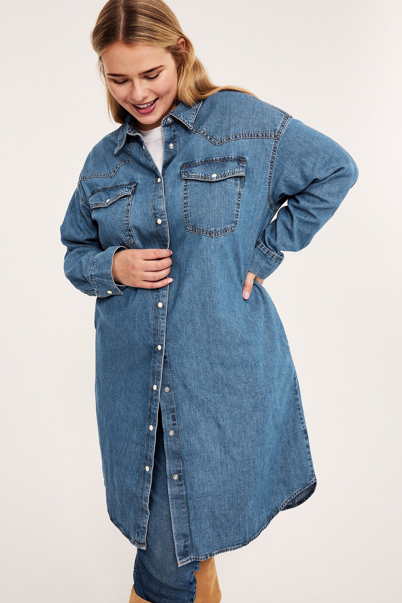 Widok z przodu kobiety w długiej niebieskiej sukience koszulowej z denimu, na białym t-shircie i jeansach.
