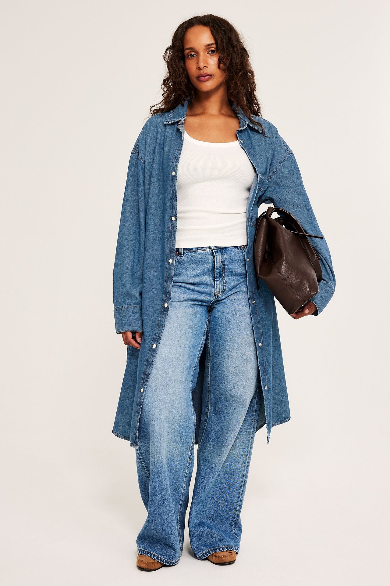 Edestä: nainen avonaisessa pitkässä sinisessä farkkupaitamekossa, valkoisessa topissa, farkuissa, ruskea laukku, kengät.