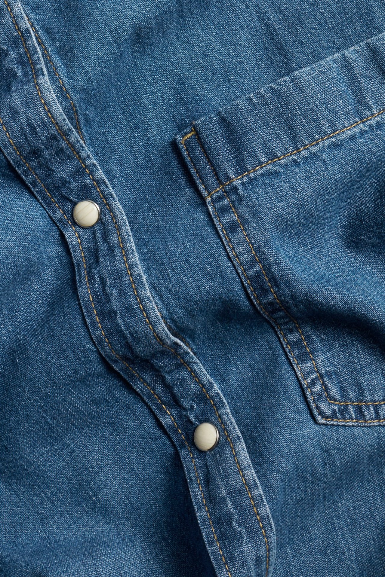 Zbliżenie na niebieską dżinsową sukienkę koszulową z jasnymi zatrzaskami, kontrastowymi szwami i kieszenią.