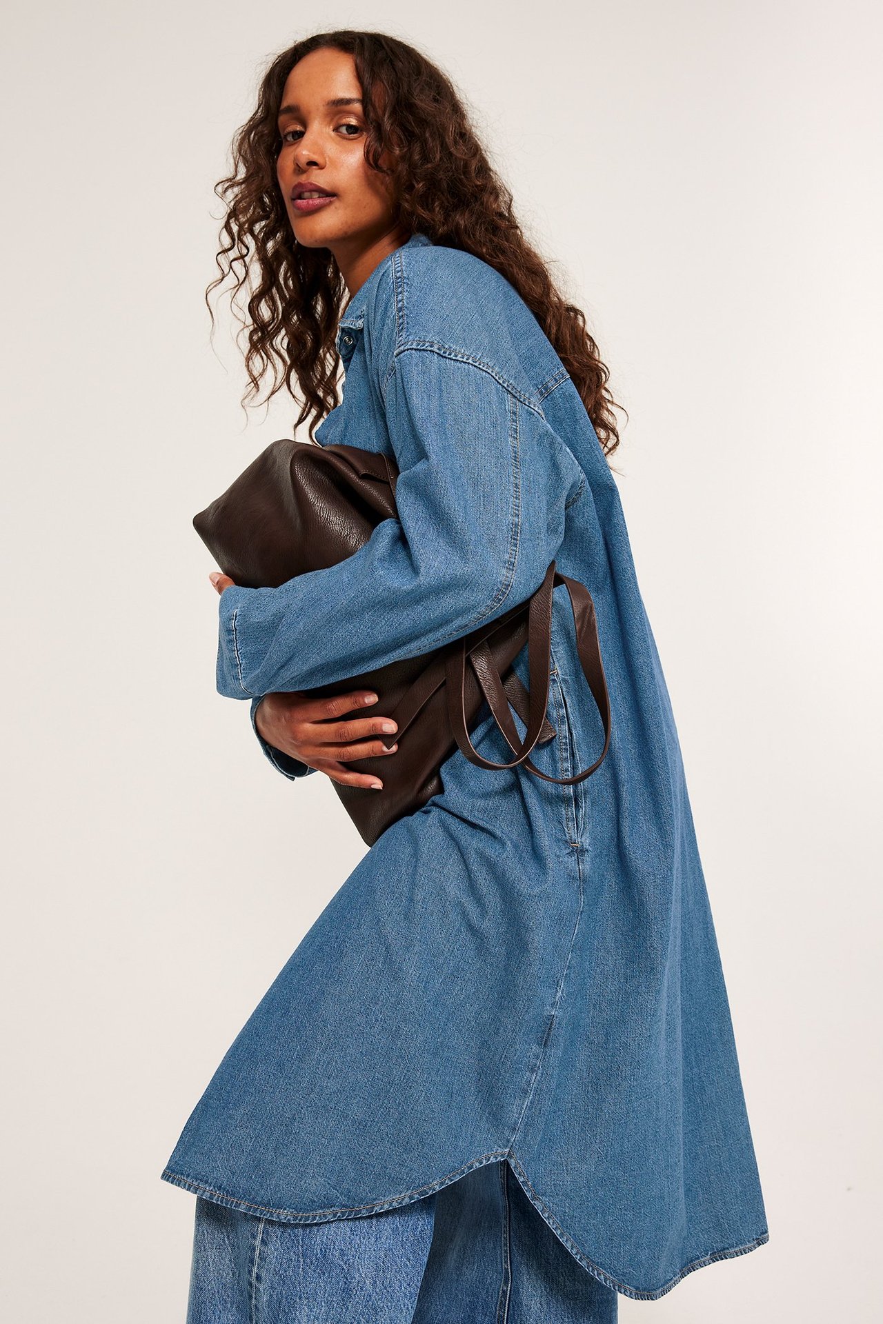 Widok z przodu: Kobieta w jasnoniebieskiej jeansowej sukience koszulowej, na jeansach, trzyma ciemnobrązową torbę.