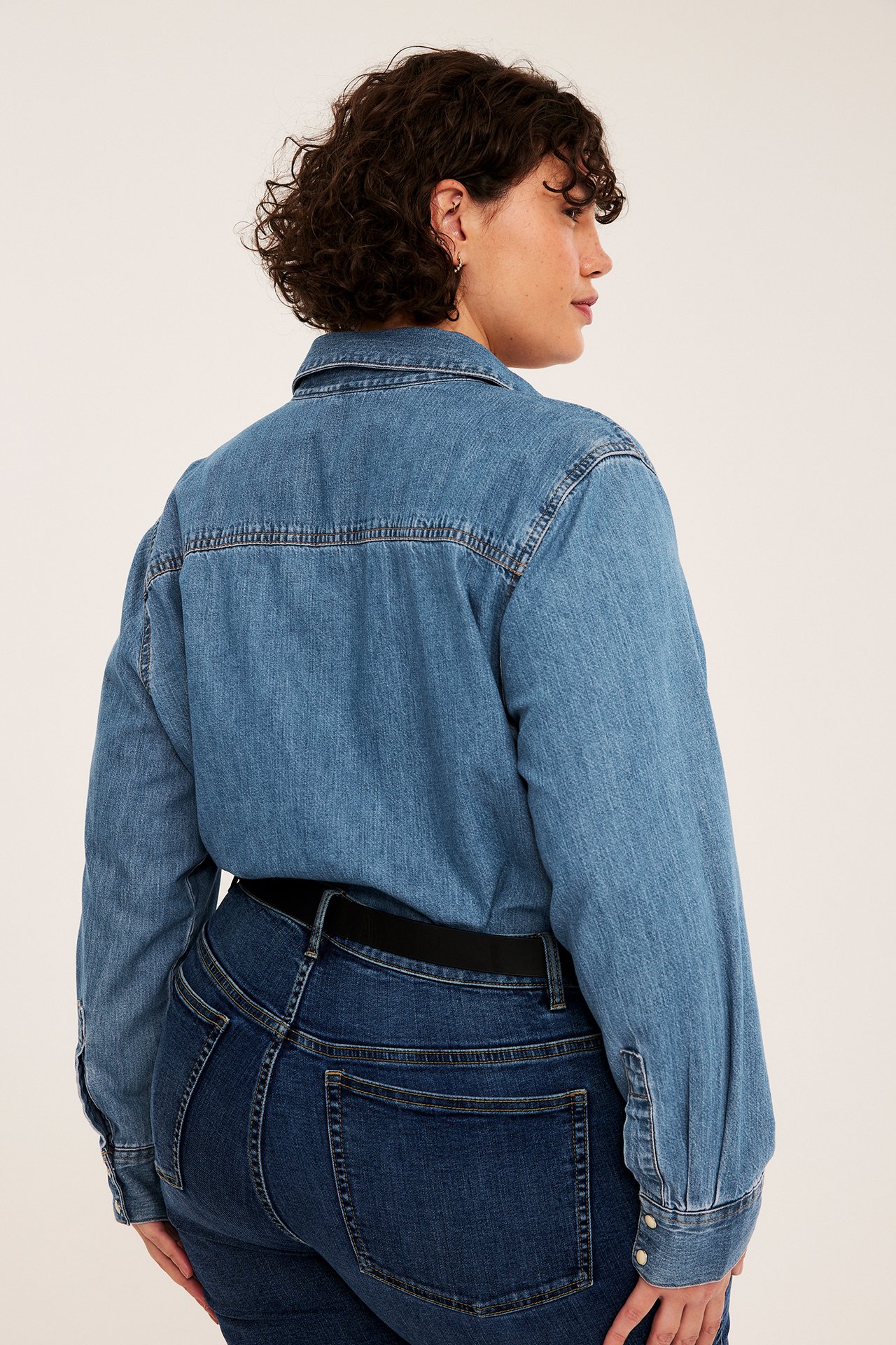 Kvinne sett bakfra, en mellomblå denimskjorte over mørke jeans med sort belte.