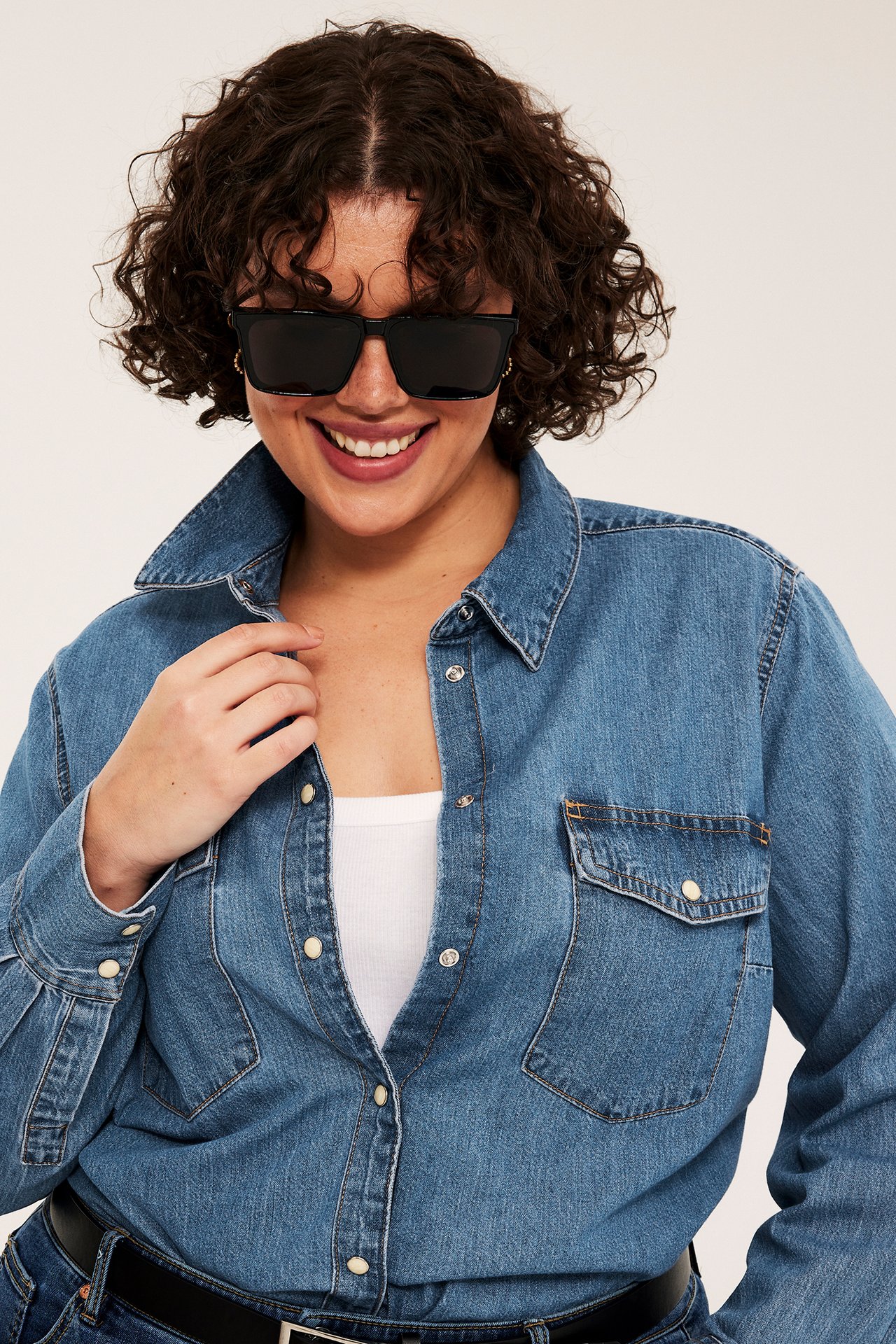 Front av kvinne i blå denimskjorte over hvit topp, svarte solbriller og svart belte.
