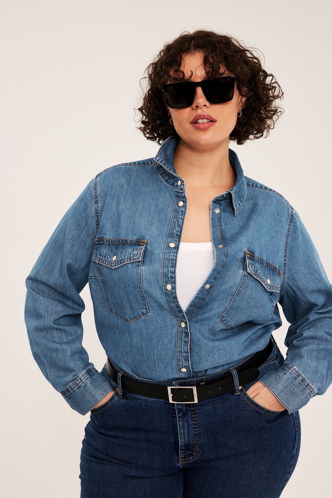 Frontbilde av kvinne i blå dongeriskjorte, mørkeblå jeans, svart belte og svarte solbriller.