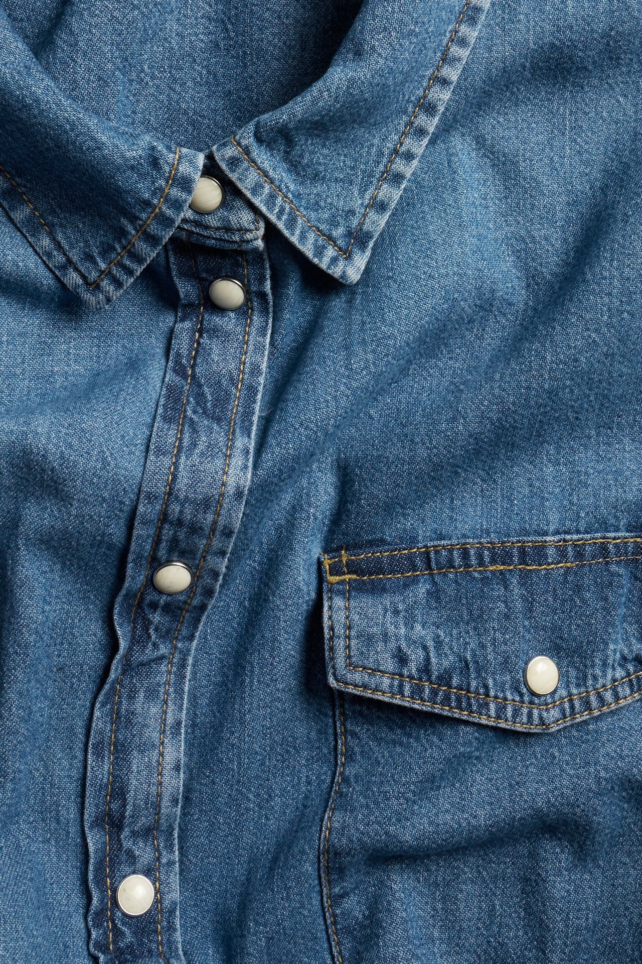 Zbliżenie niebieskiej bawełnianej koszuli jeansowej z białymi zatrzaskami i kieszenią na piersi.