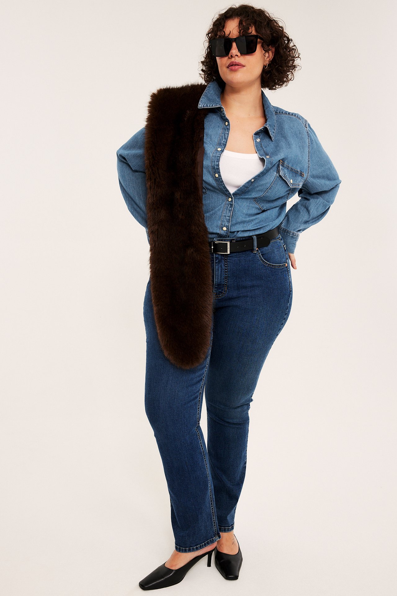 Kvinne i mørkebrun fuskepelsstola, blå denimskjorte, blå jeans og svarte mules, forfra.