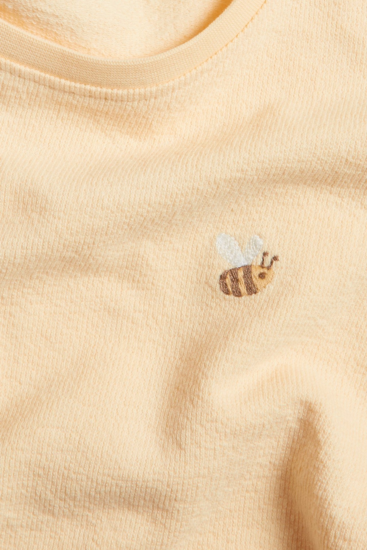 Närbild på en gul bomulls-t-shirt med en liten broderad bi på framsidan.