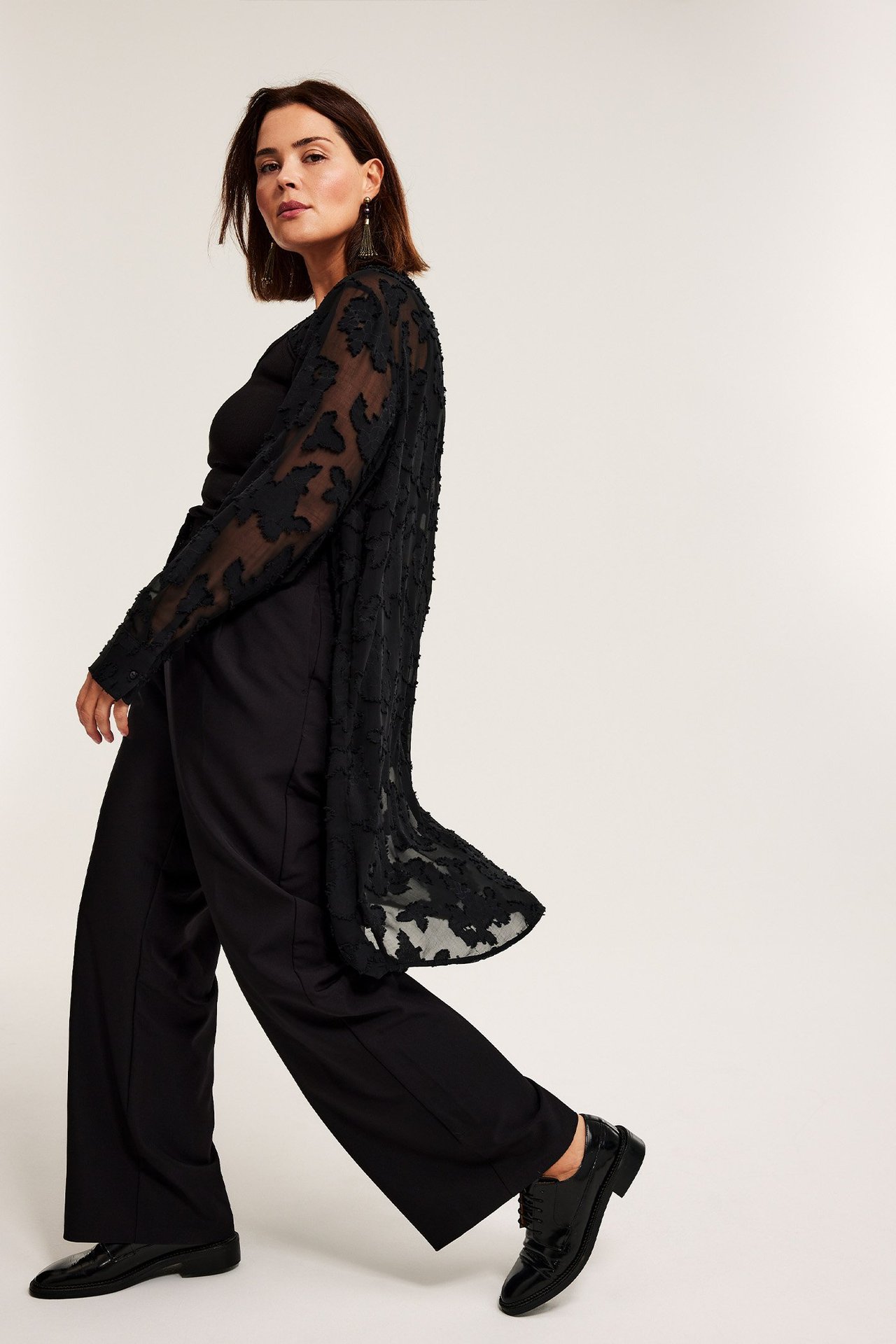 Sidesyn: Kvinne i svarte vide dressbukser, en svart topp og en mønstret lang svart cardigan.