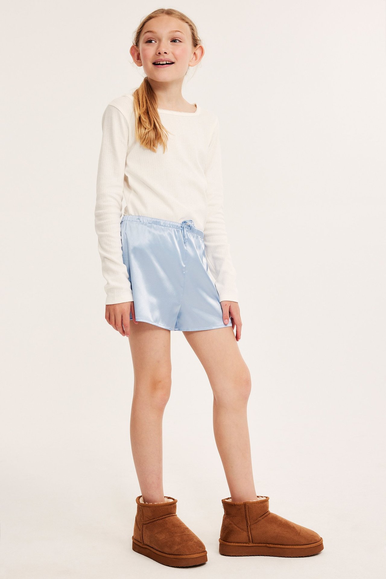 Barn tjej i vit långärmad tröja och ljusblå satin pyjamasbyxor med dragsko, frontvy.