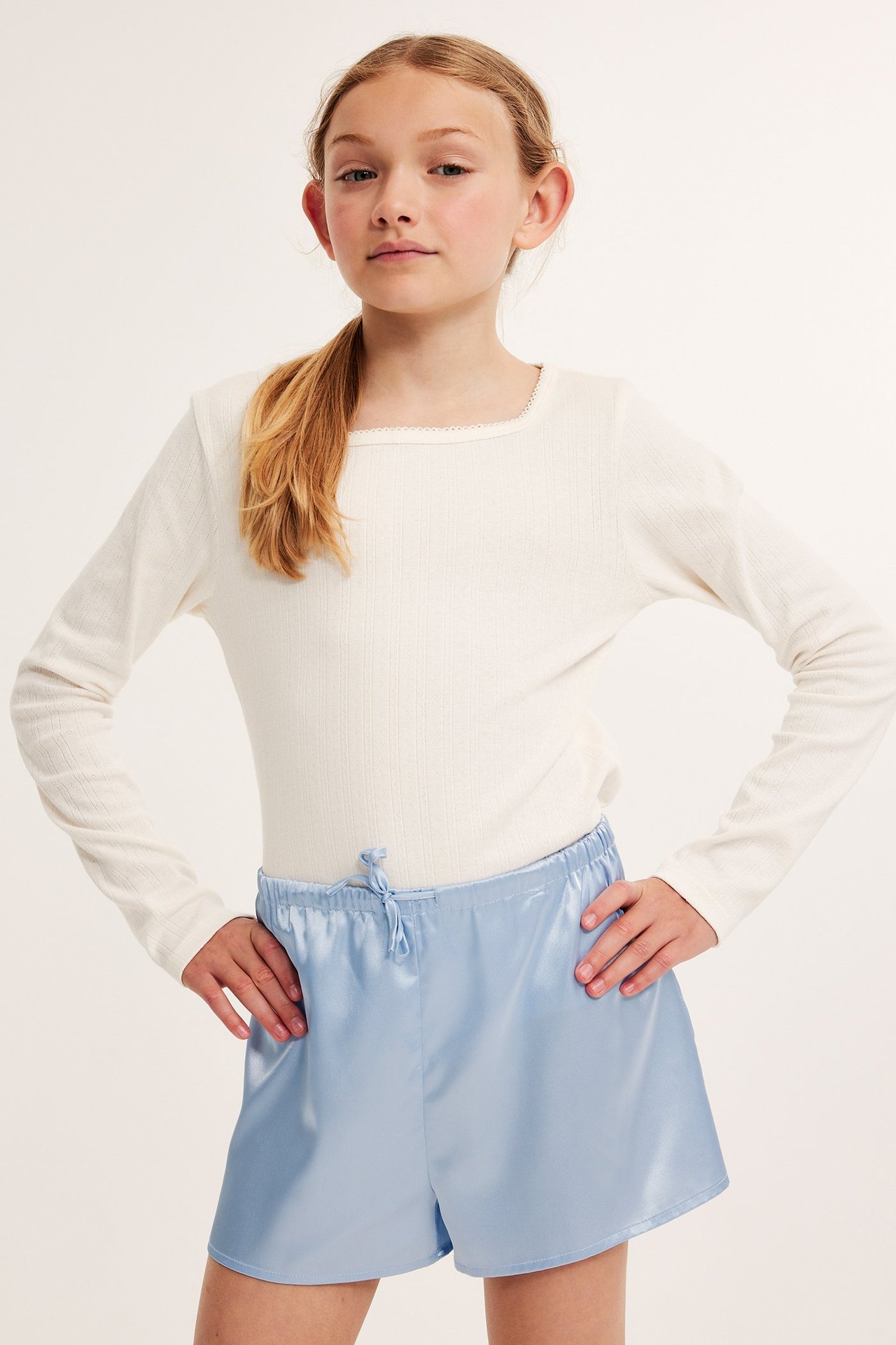 Kvinna i vit långärmad tröja och ljusblå satinpyjamasbyxor med dragsko i midjan.