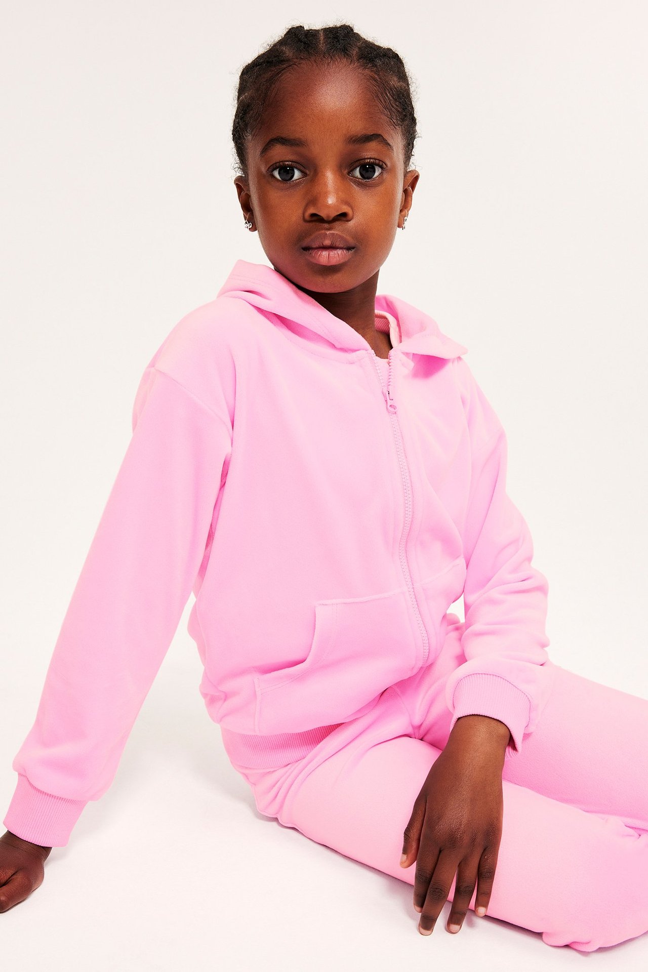 Framifrån: ett barn i ljusrosa velourhoodie med dragkedja, fickor och matchande byxor.