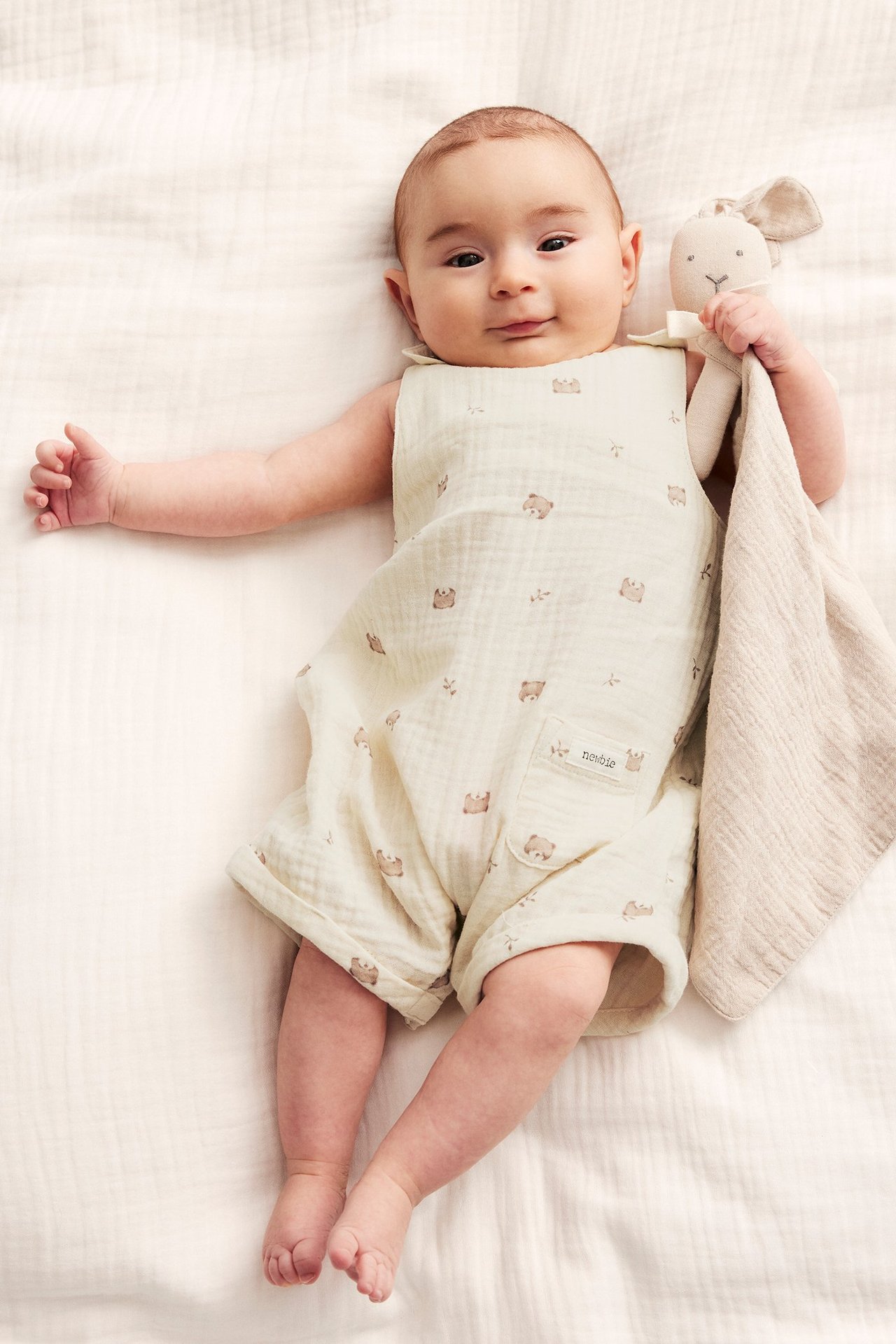 Uppifrån sett, baby i ljusbeige bomulls-playsuit med nallarmönster, håller en kaninsnuttis.