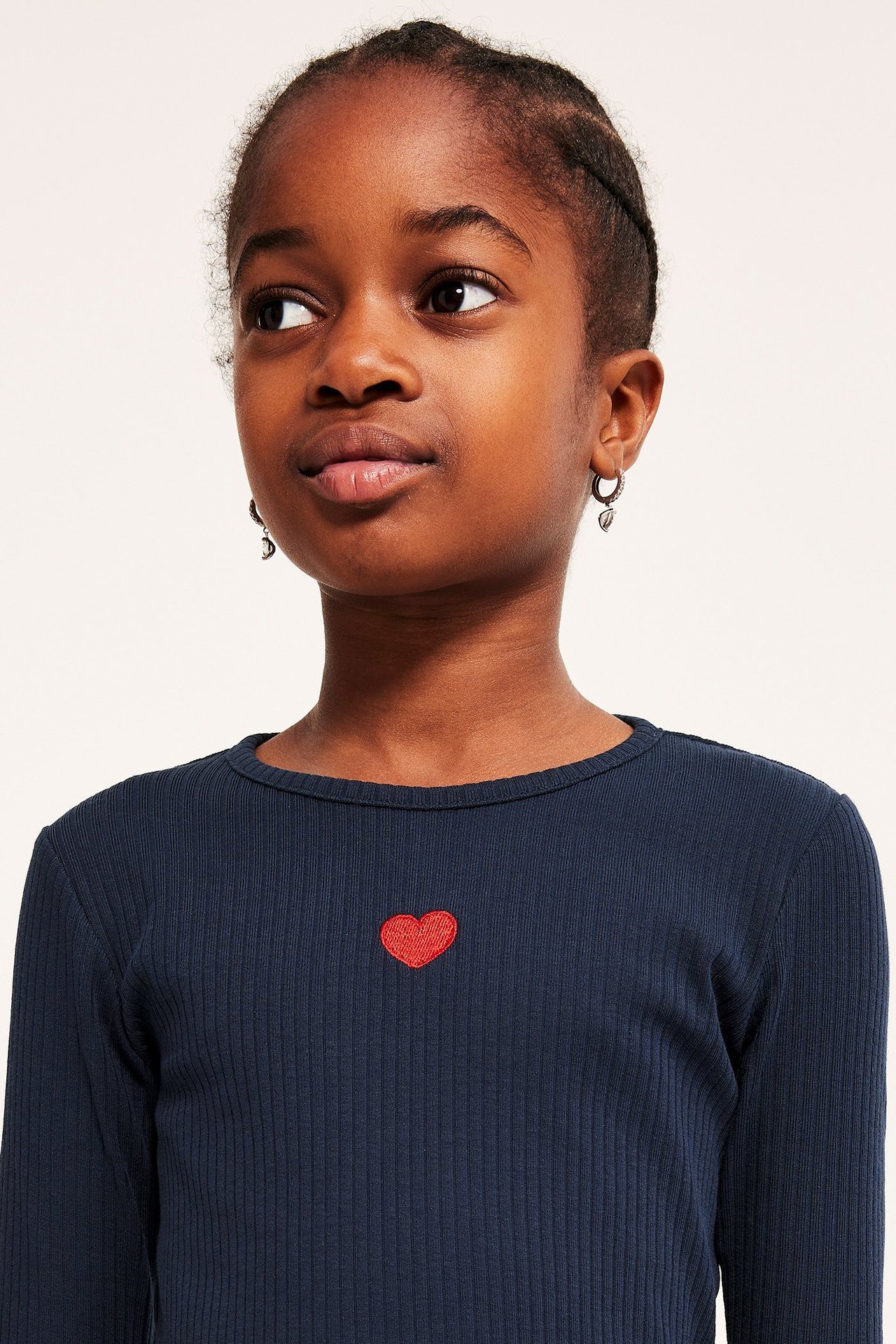 Dziecko widziane z przodu. Dziecko odwraca wzrok od obrazu i ma krótkie, ciemne, splecione włosy. Ubrane jest w ciemnoniebieską koszulkę z długim rękawem, z czerwonym serduszkiem na piersi i małymi, zwisającymi kolczykami.