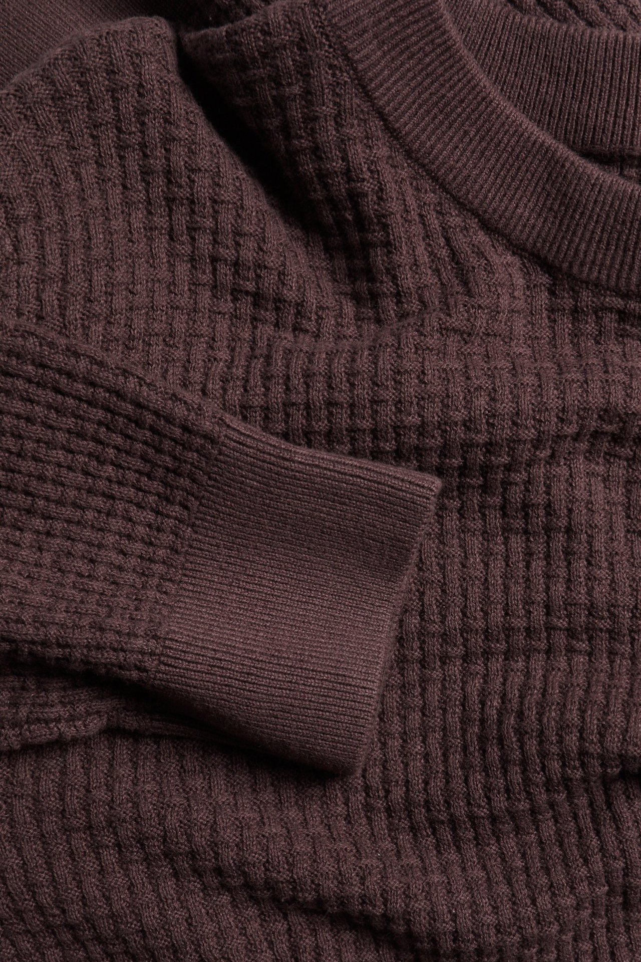 Zbliżenie ciemnofioletowego bawełnianego swetra z dzianiny o wzorzystej fakturze, ściągaczach i okrągłym dekolcie.