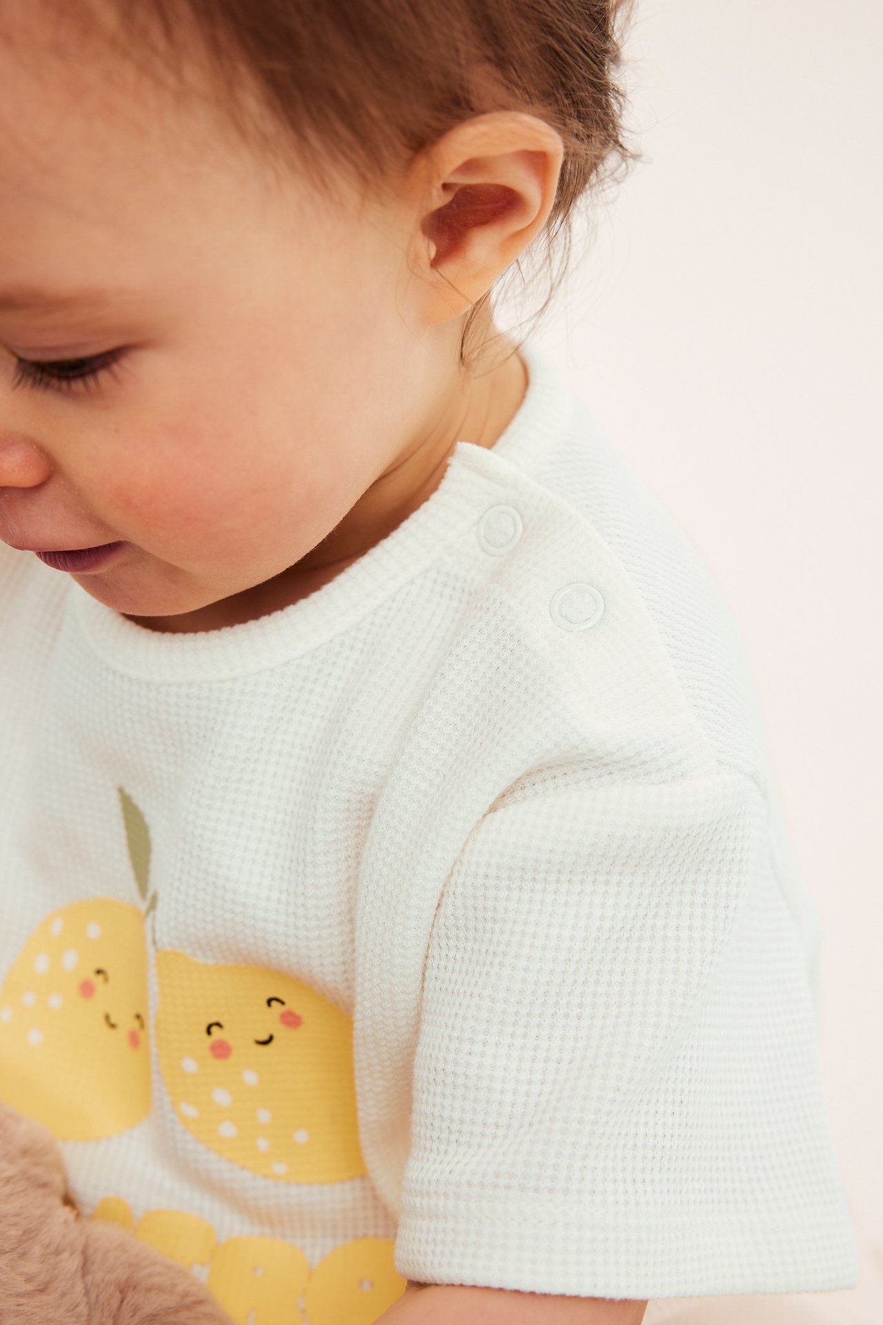 Nærbilde, sidevisning av baby i offwhite vaffelstrikket t-skjorte med gult sitronmønster og trykknapper.