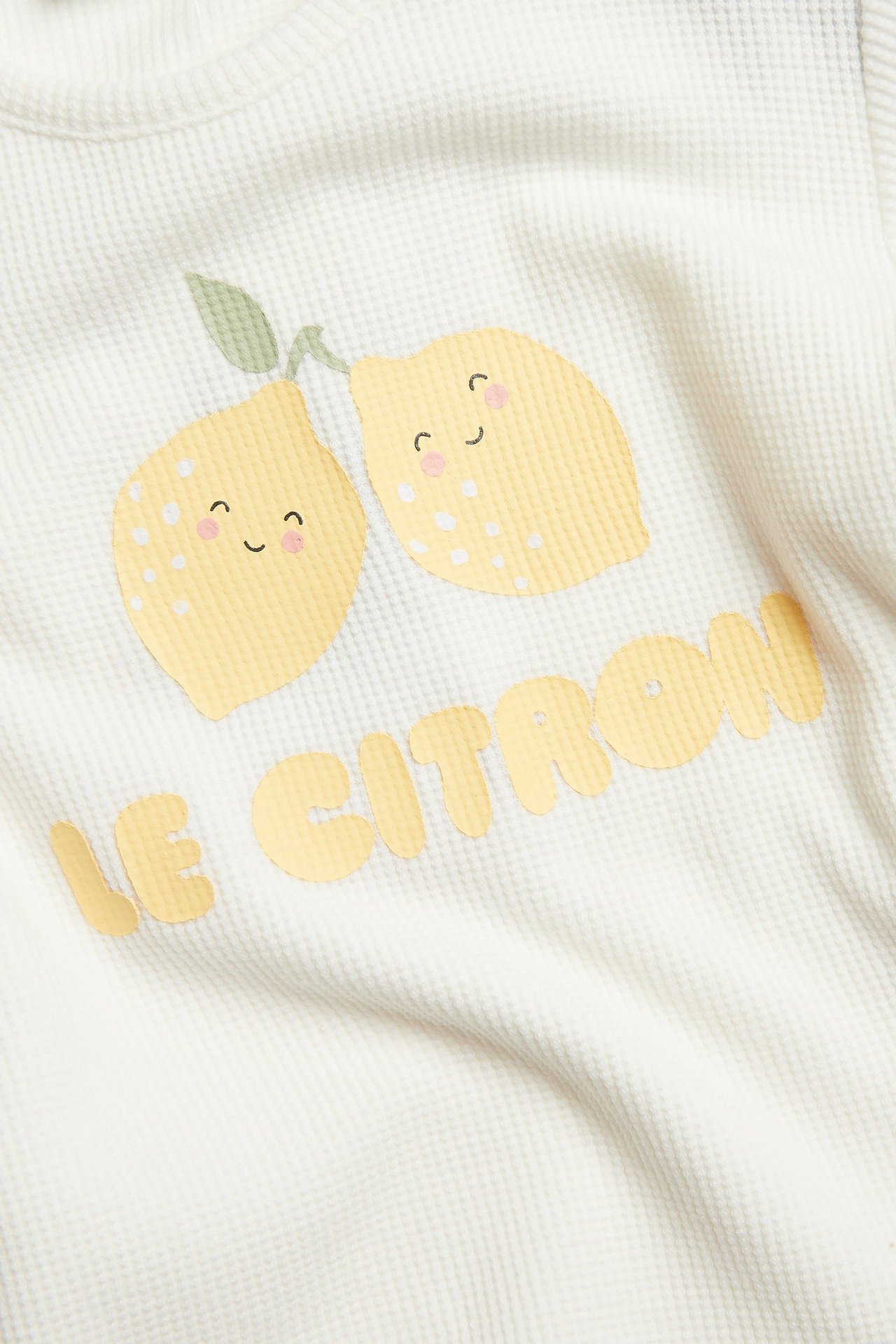 Zbliżenie na ecru waflowy T-shirt niemowlęcy z żółtym nadrukiem cytryn i napisem 'LE CITRON'.