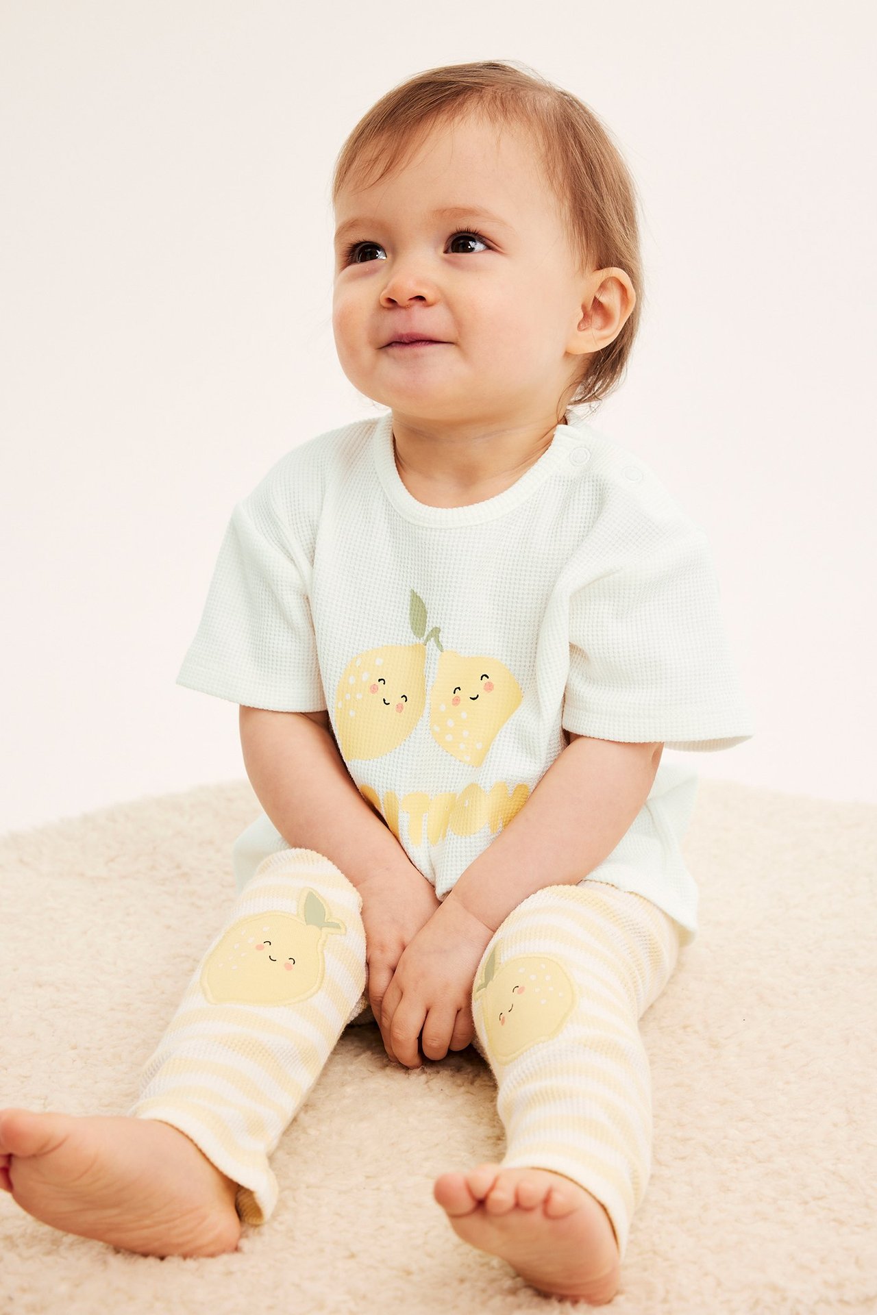 Widok z przodu: niemowlę w ecru gofrowanym T-shircie z nadrukiem cytryn, napami i pasiastych legginsach.