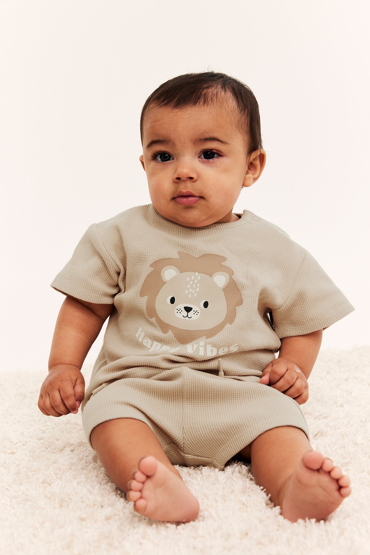 Frontvy av en baby i beige våfflad kortärmad romper med lejonprint framtill.