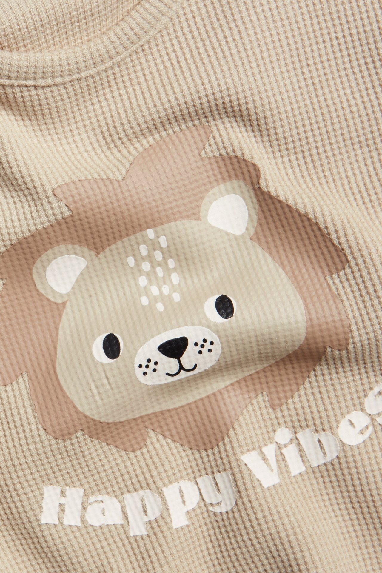 Närbild framifrån på en beige våfflad baby-t-shirt med lejonhuvudtryck och texten 'Happy Vibes'.