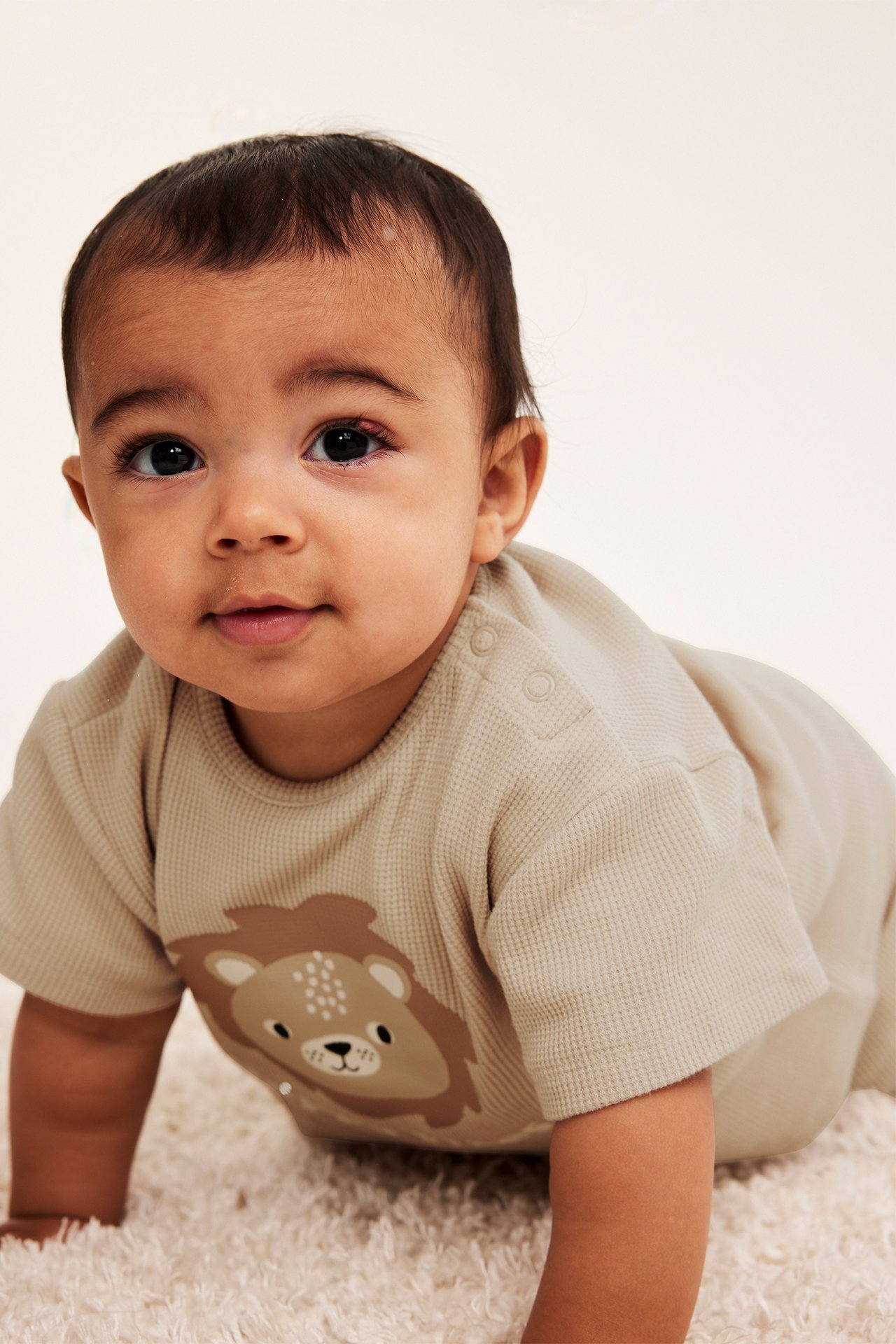 Frontvy av en baby i en beige våfflad t-shirt med lejontryck och tryckknappar på axeln.