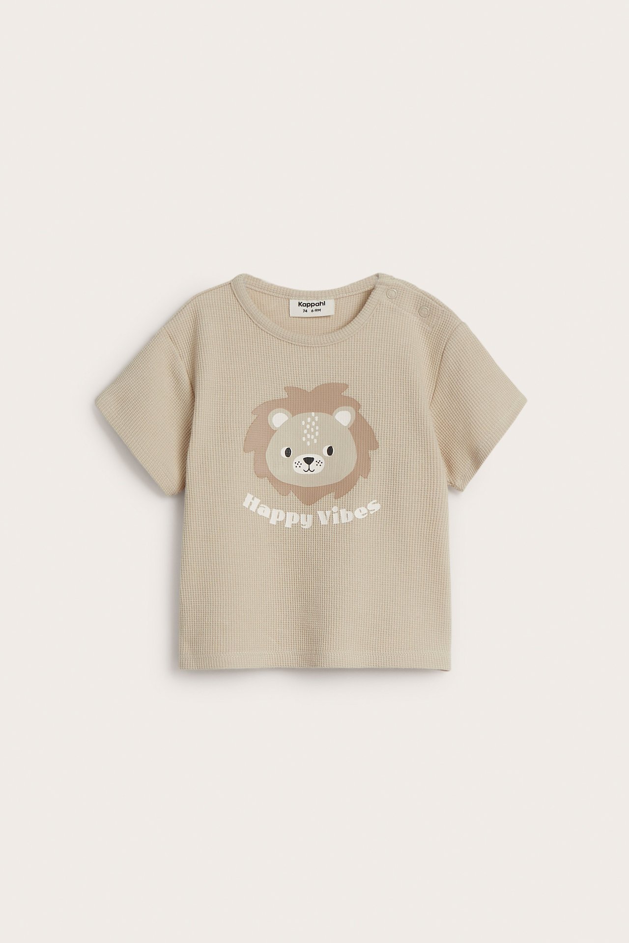 Frontvy av en beige våfflad baby-t-shirt med lejonansikte och tryckknappar på axeln.