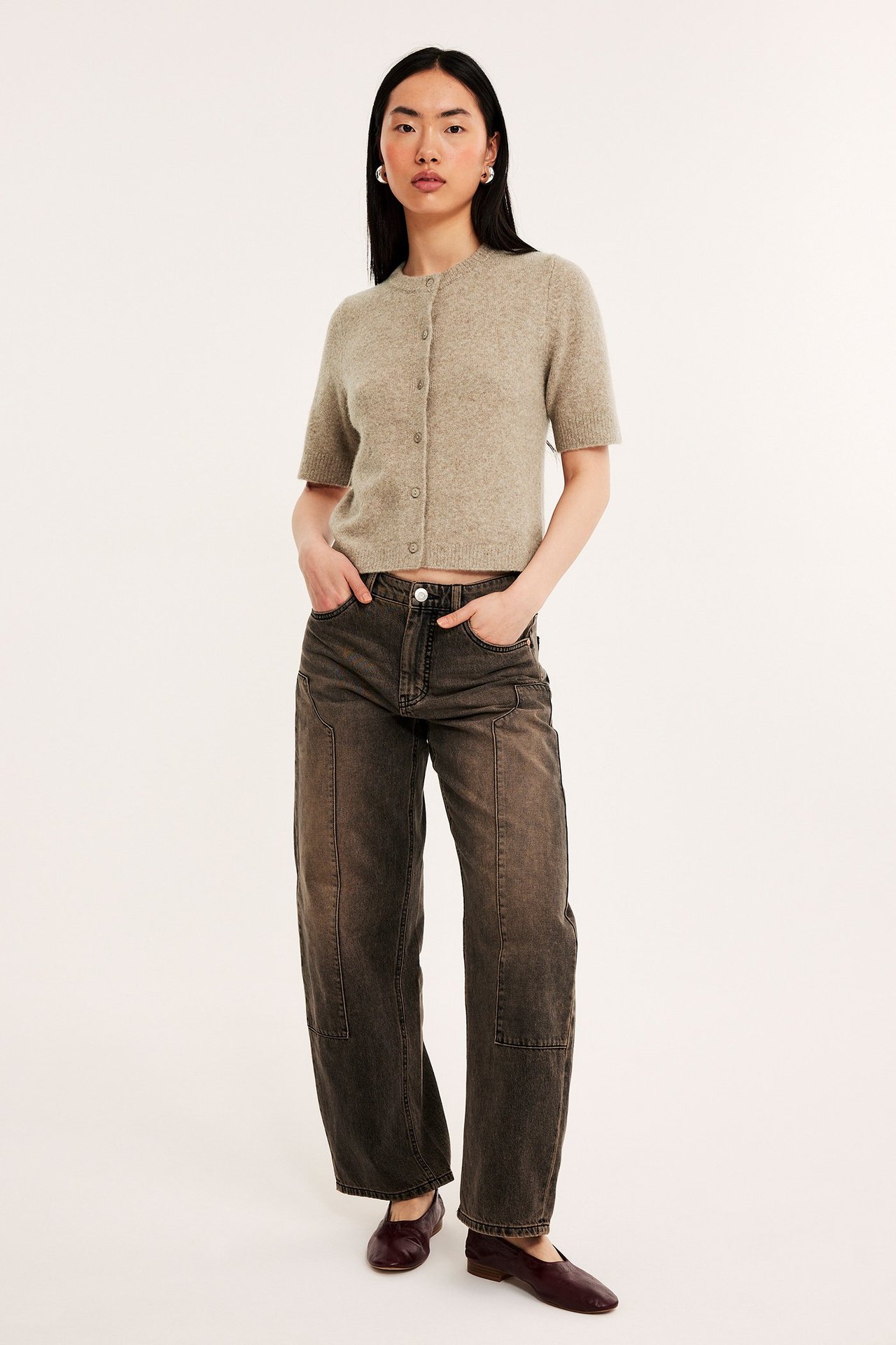 Frontbilde av kvinne i lys brunmelert kortermet strikket cardigan, mørke vide bukser og burgunder flate sko.