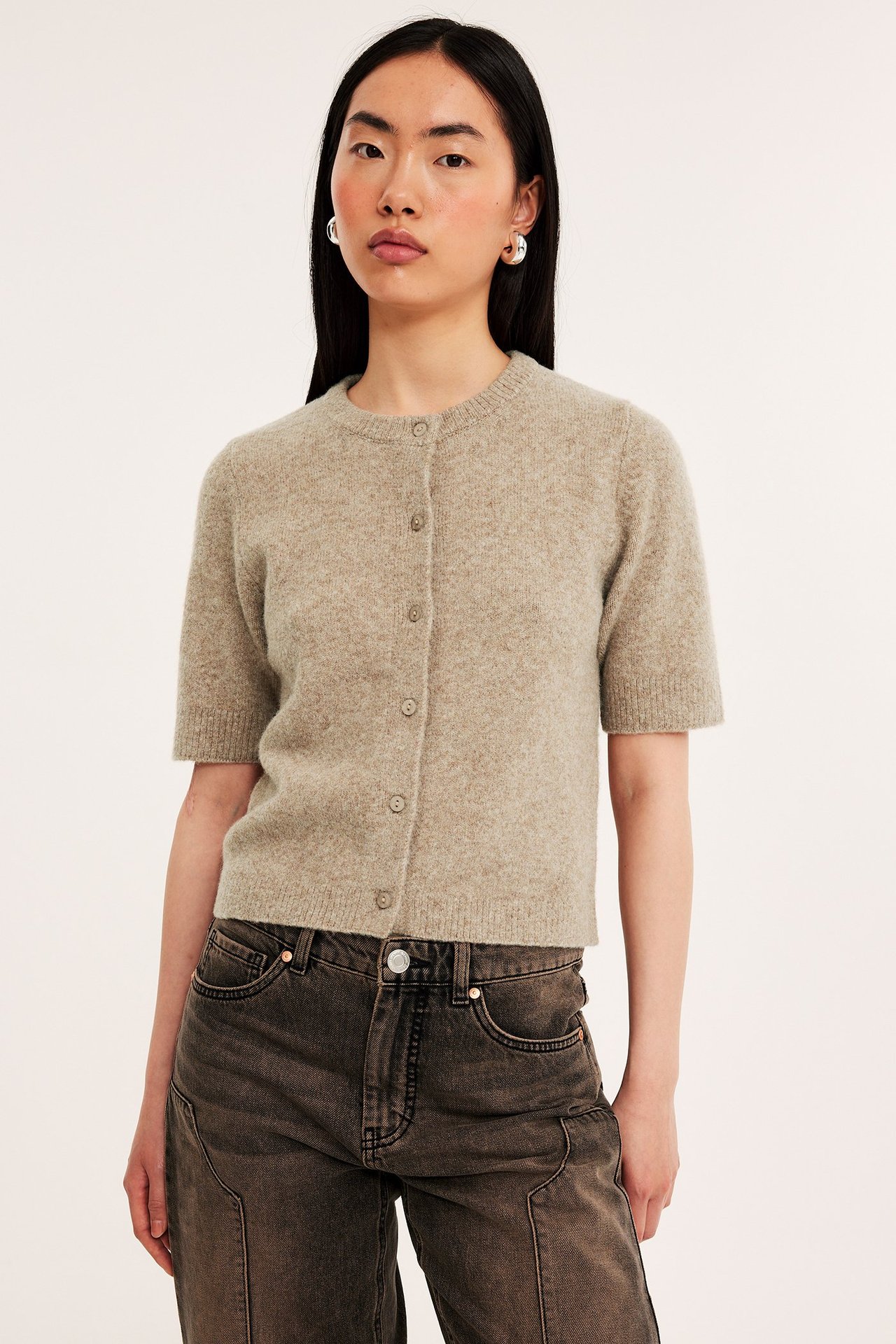 Forfra: kvinne i melert beigebrun kortermet strikket cardigan, mørke jeans og sølvfargede øreringer.