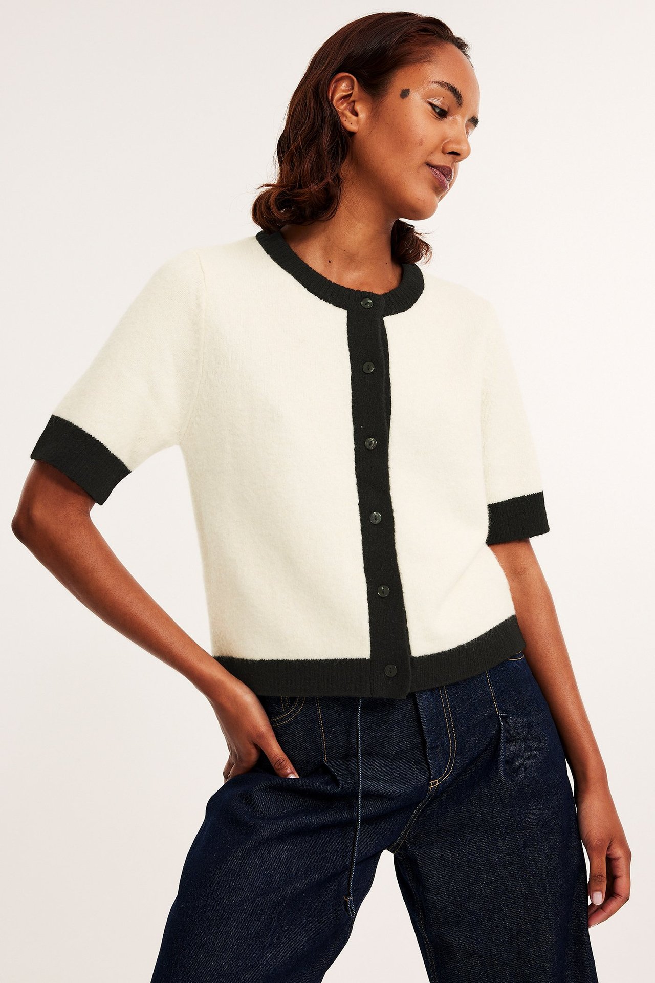 Forfra: kvinne i offwhite kortermet strikket cardigan med svarte detaljer og mørkeblå jeans.