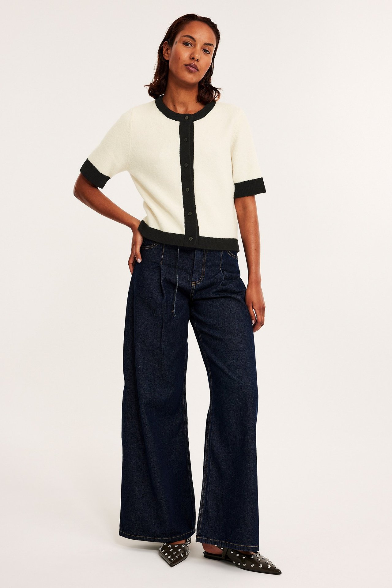 Kvinne i offwhite kortermet strikket cardigan med svart kant, mørkeblå vide bukser og svarte nagle-mules.