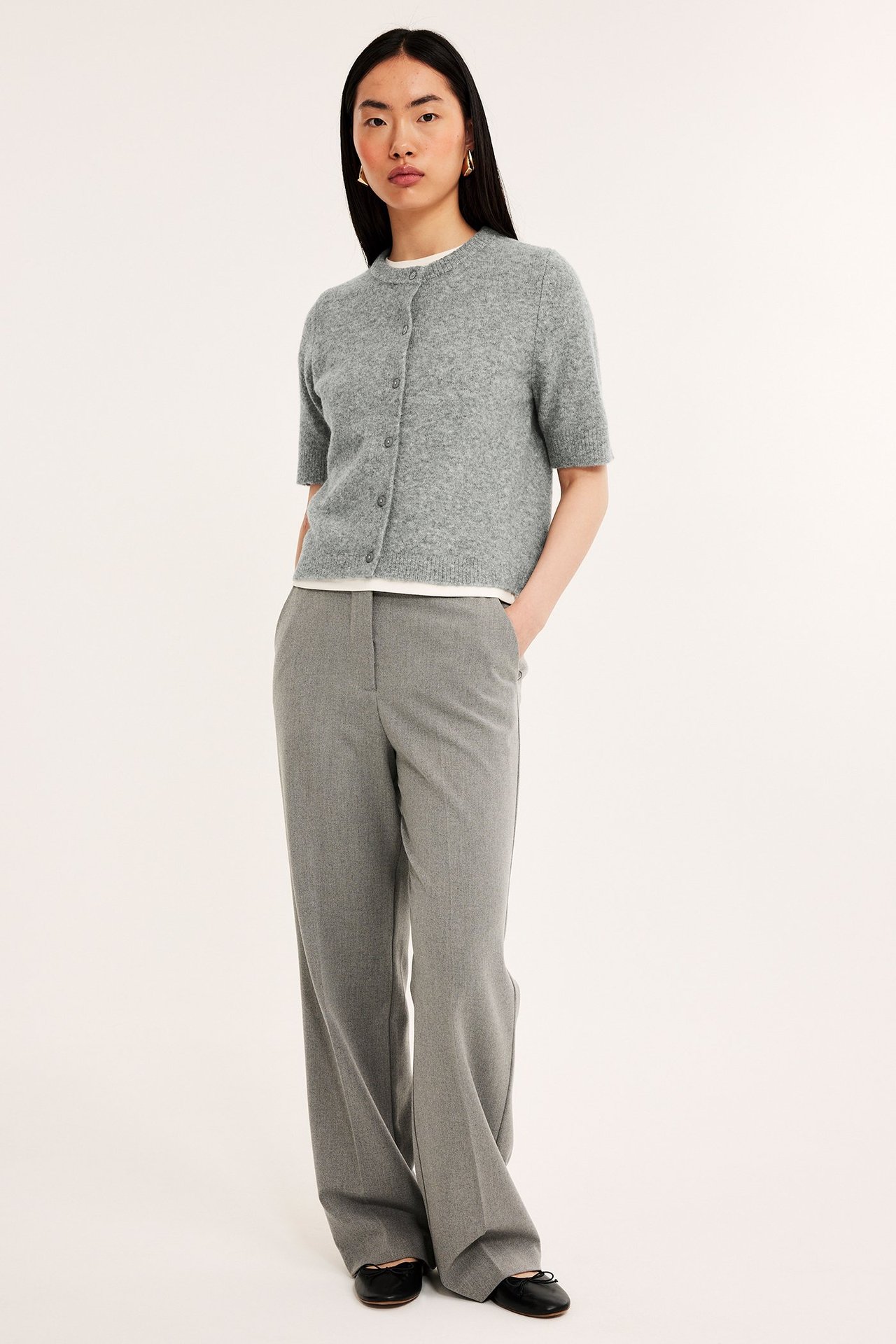 Front: kvinne i lys grå melert kortermet strikket cardigan over hvit t-skjorte, grå bukser, svarte ballerinasko.