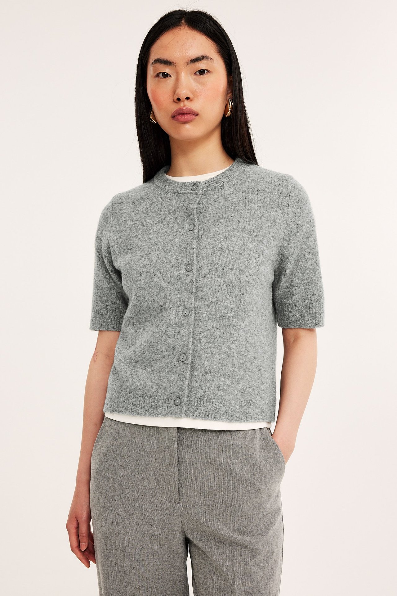 Forfra: kvinne i lys grå melert kortermet strikket cardigan over hvit t-skjorte og grå bukser.