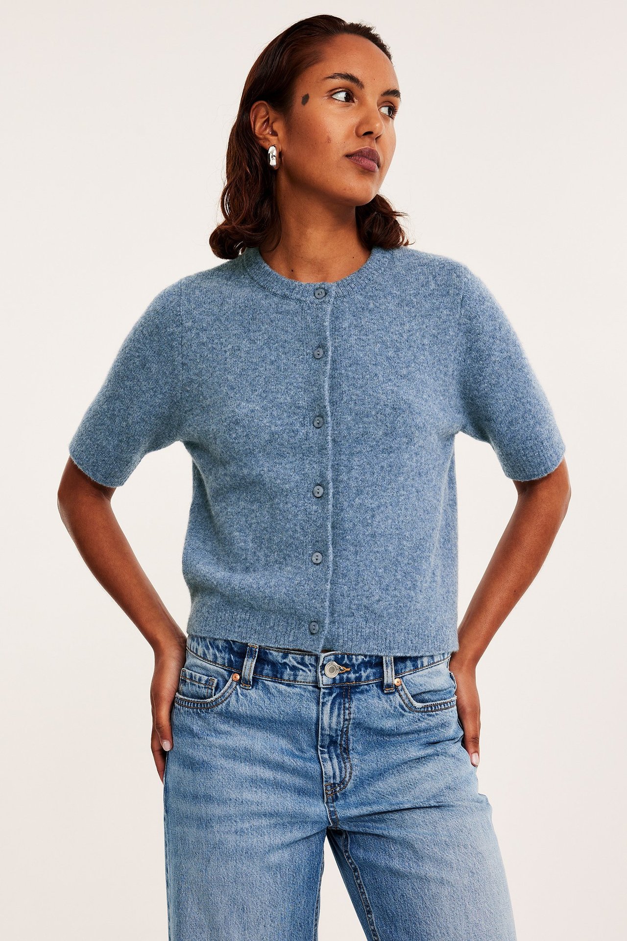 Edestä: nainen yllään vaaleansininen meleerattu lyhythihainen neulejakku, siniset farkut ja hopeiset korvarenkaat.