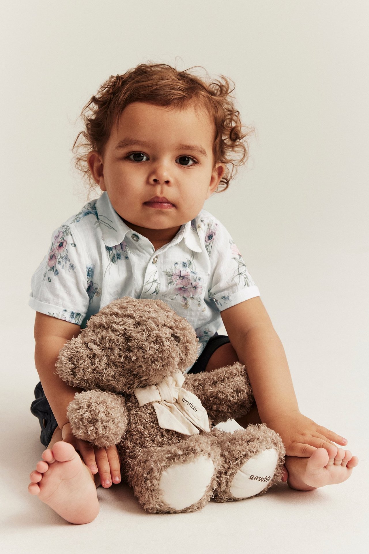Widok z przodu: niemowlę w kremowej kwiecistej koszuli z krótkim rękawem, ciemnych szortach, trzyma brązowego misia.