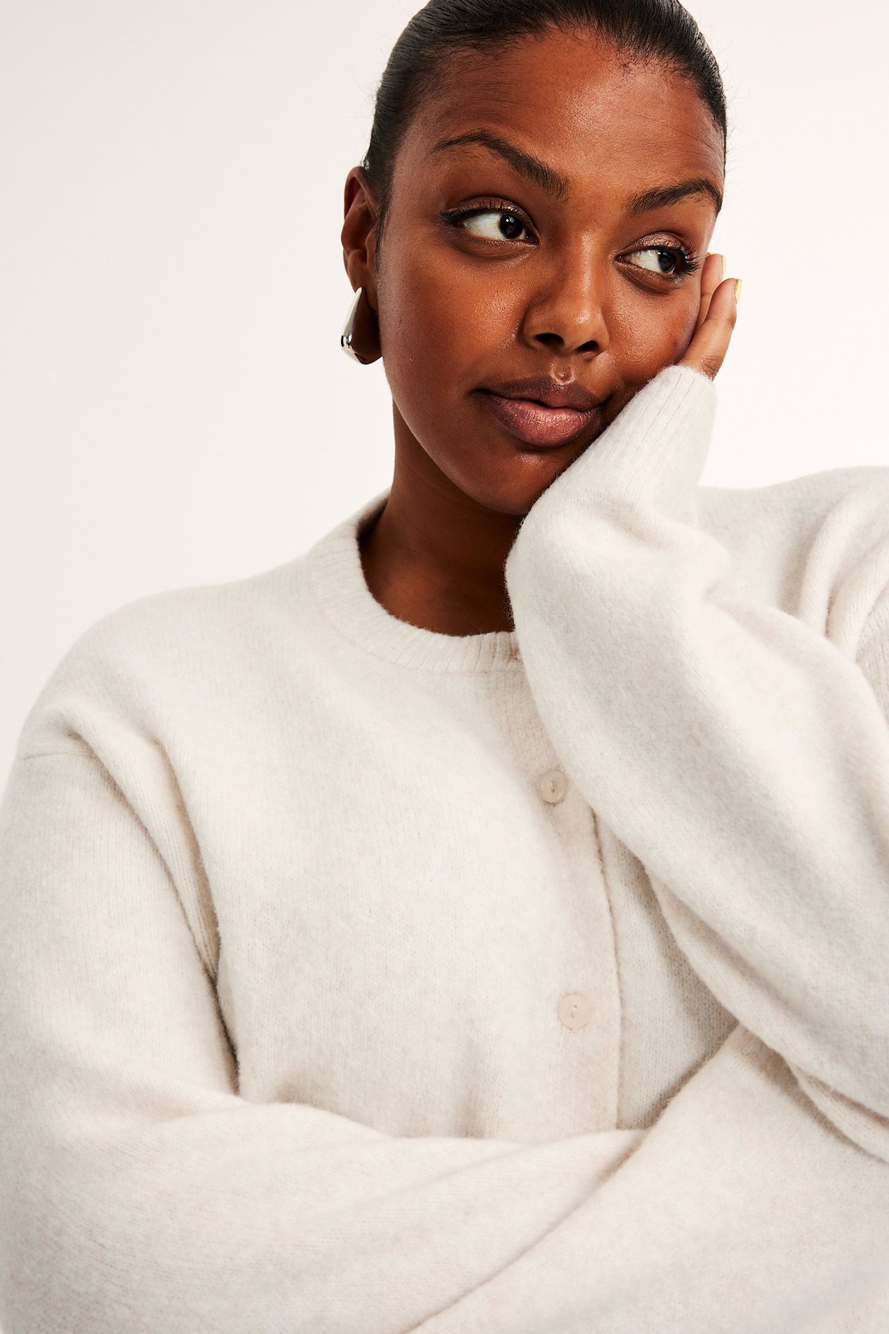 Nærbilde av en kvinne i en lys beige finstrikket cardigan med knapper og gullfargede øreringer.