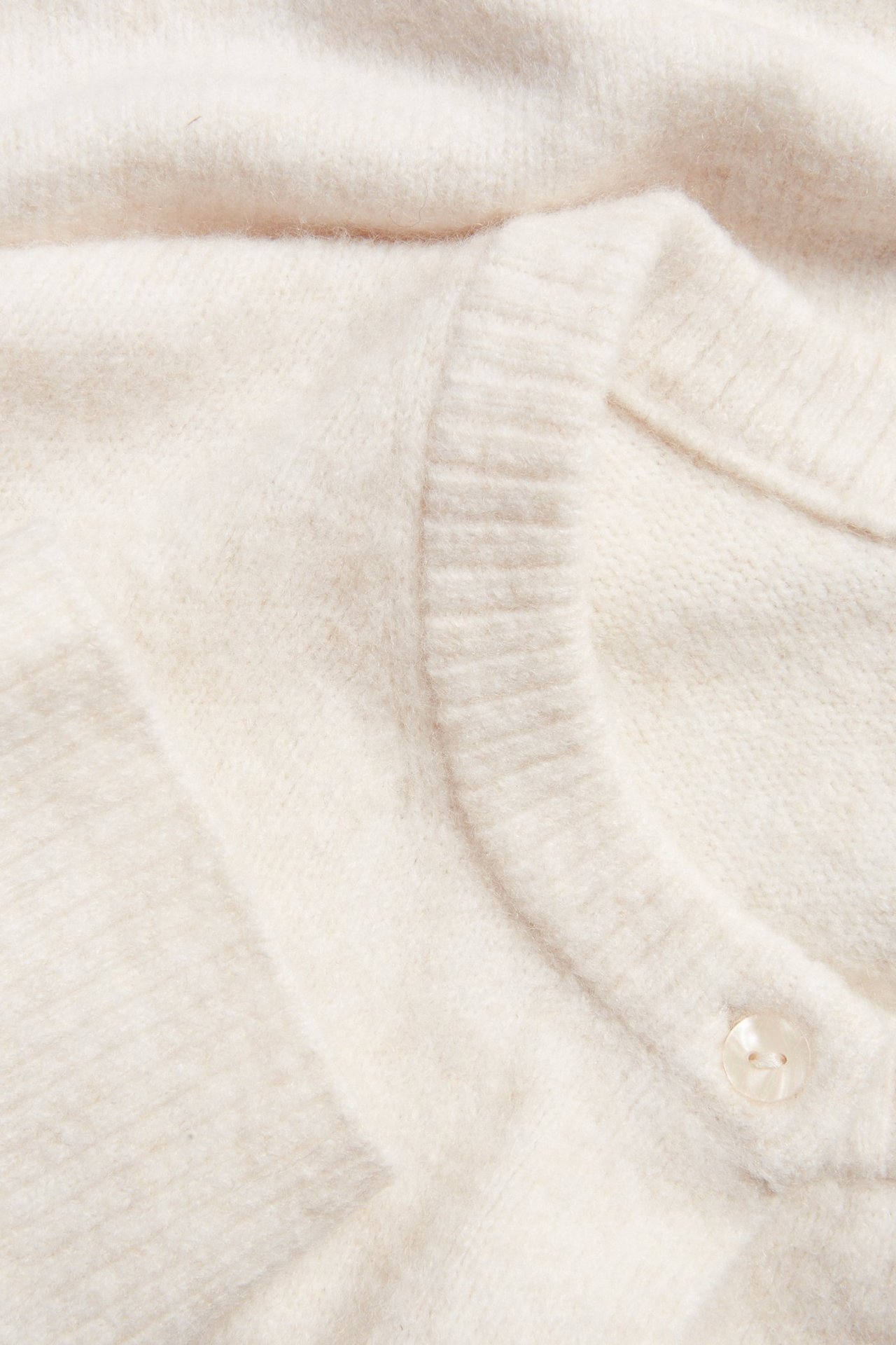 Zbliżenie jasnobeżowego, drobno dzianego kardiganu z okrągłym dekoltem i jednym widocznym guzikiem.