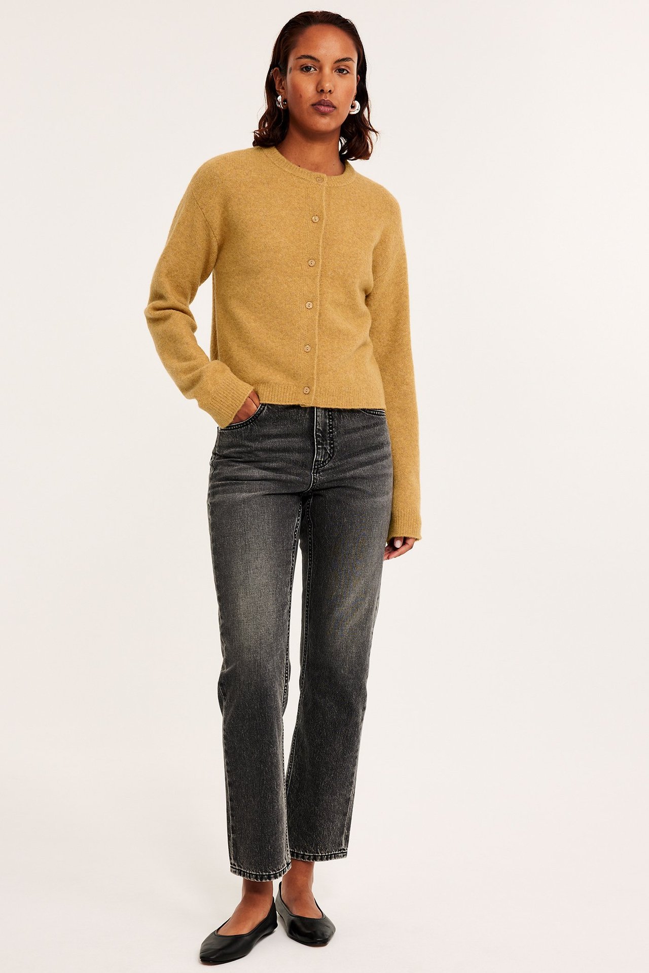 Nainen yllään keltainen hienoneulottu napitettu neuletakki, tummanharmaat farkut, mustat ballerinat ja hopeakorvakorut.