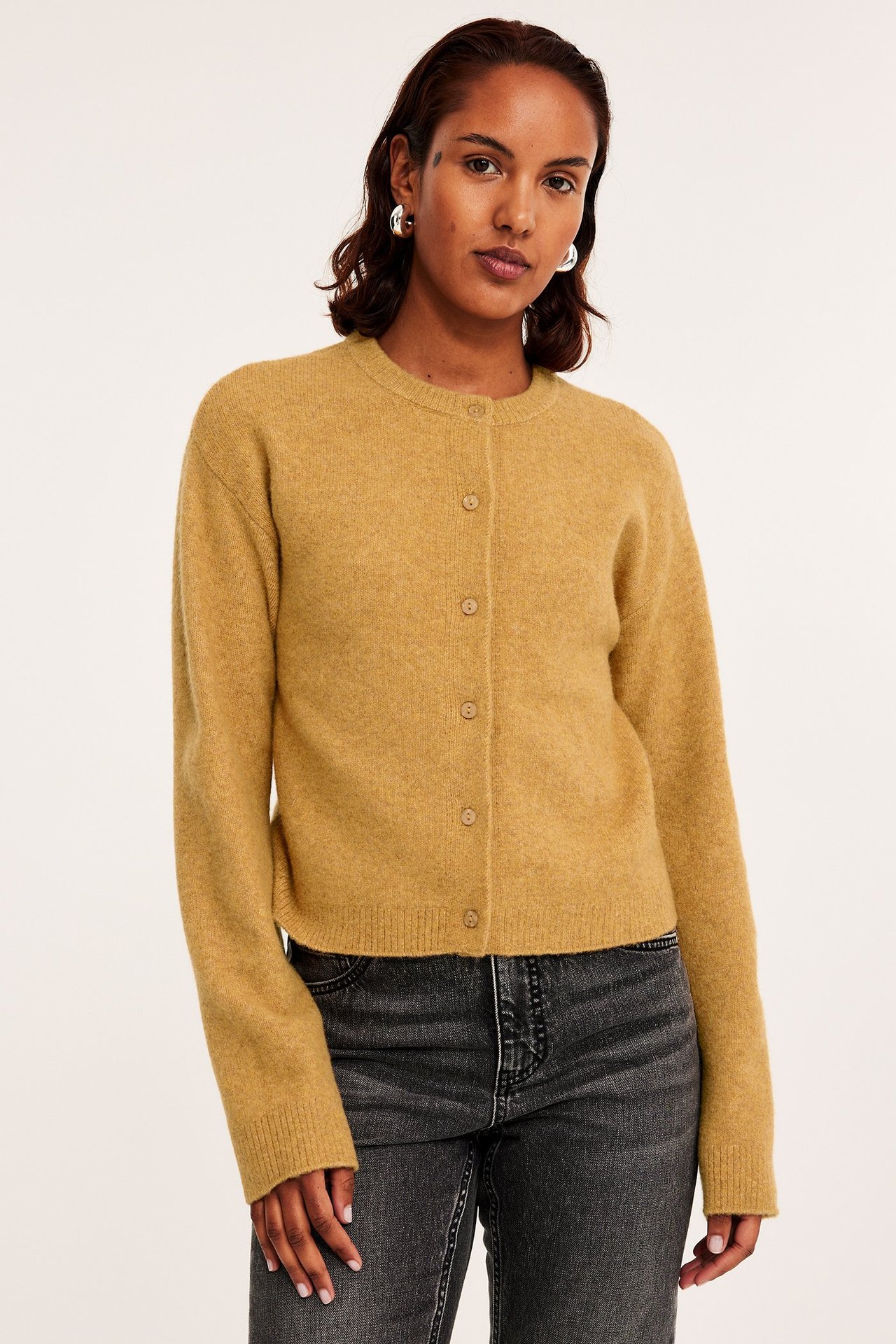 Edestäpäin, nainen yllään sinapinkeltainen hienoneulottu napillinen neuletakki ja tummanharmaat farkut.