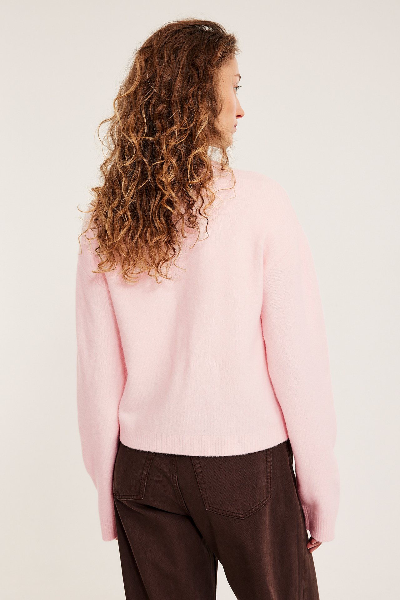 Widok z tyłu kobiety w jasnoróżowym, drobno dzianym kardiganie i ciemnobrązowych spodniach.