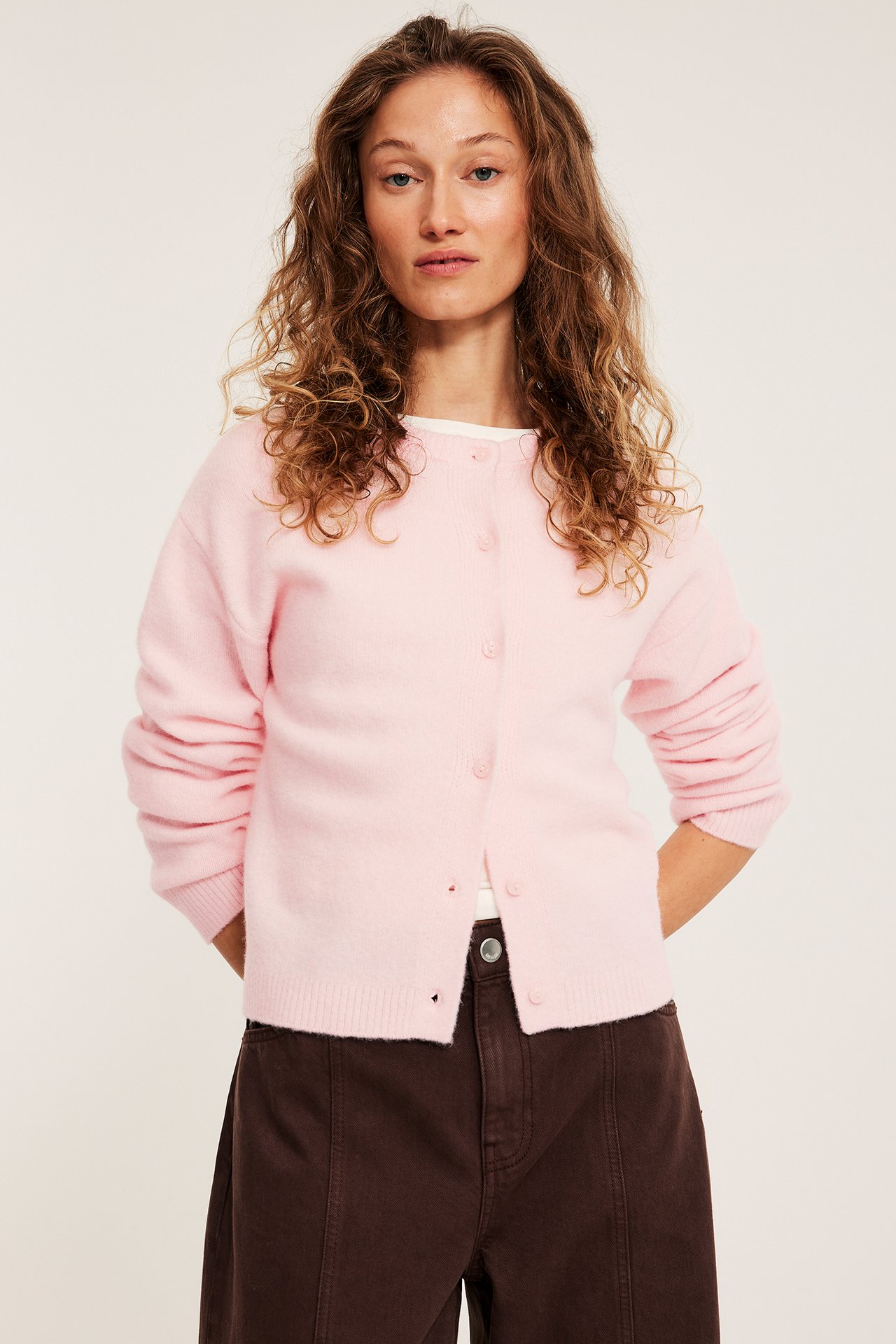 Widok z przodu: Kobieta w jasnoróżowym, drobno dzianym zapinanym swetrze, białym topie i ciemnobrązowych spodniach.
