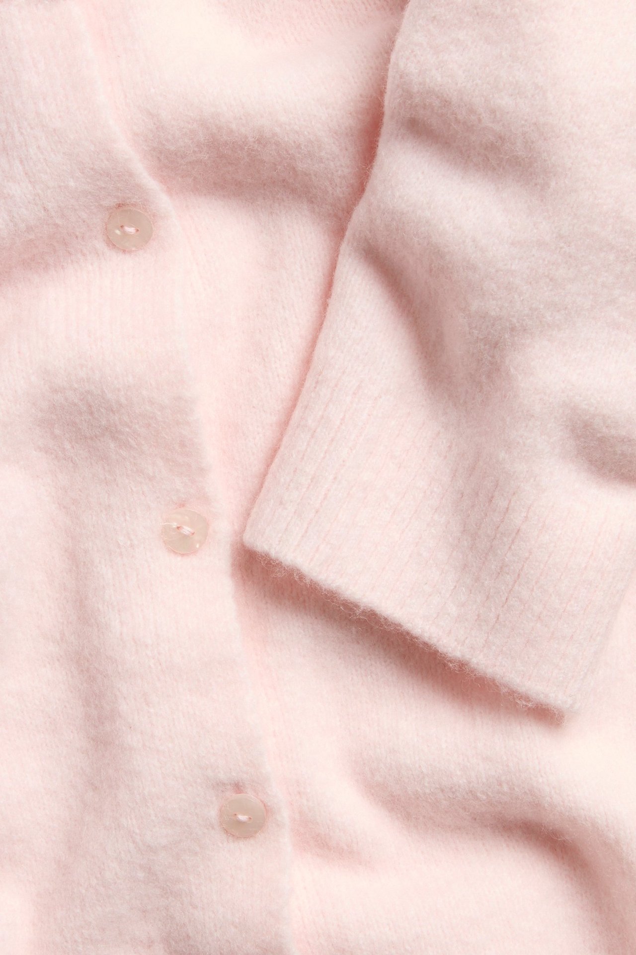 Nærbilde av en lys rosa finstrikket cardigan med tre synlige knapper og ribbestrikket mansjett.