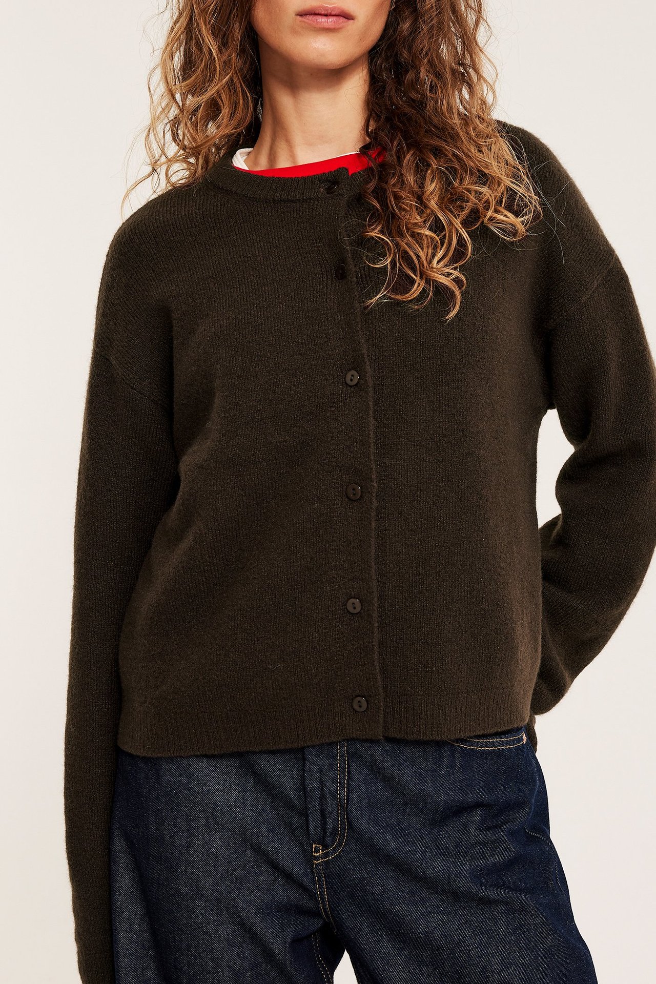 Kvinna i en mörkbrun finstickad kofta med knappar över en vit/röd topp och mörkblå jeans.