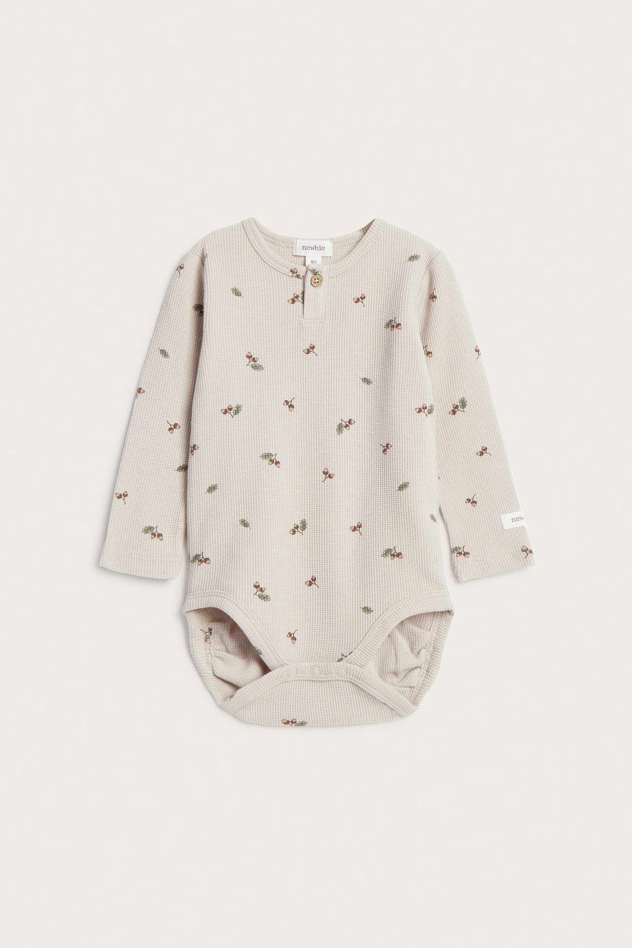 Frontvisning av en beige vaffelstrikket babybody med lange ermer, eikenøttmønster og knapper foran.