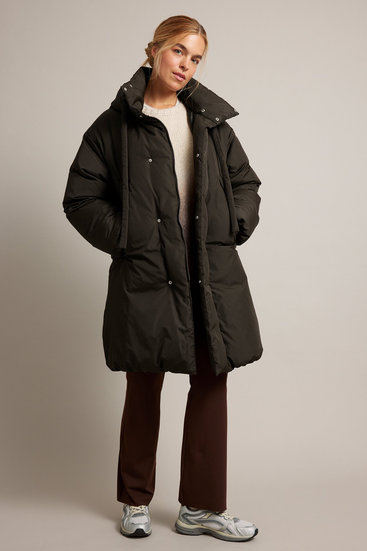 Kvinnlig modell i mörkgrön lång puffjacka över beige tröja, bruna byxor och sneakers. Frontvy.
