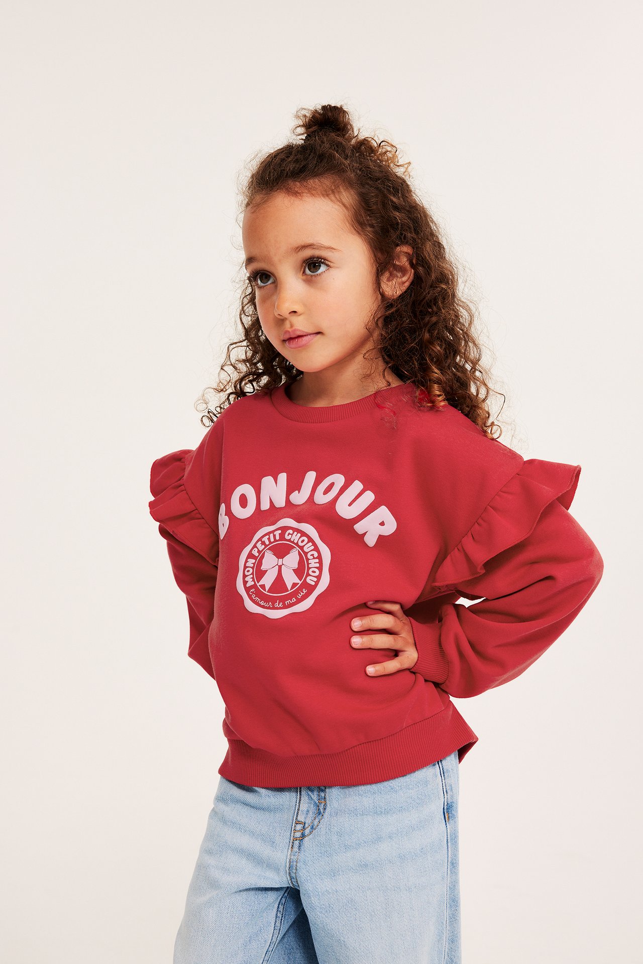 Framifrån: Ett barn i röd sweatshirt med volanger och "BONJOUR"-tryck, matchat med ljusblå jeans.