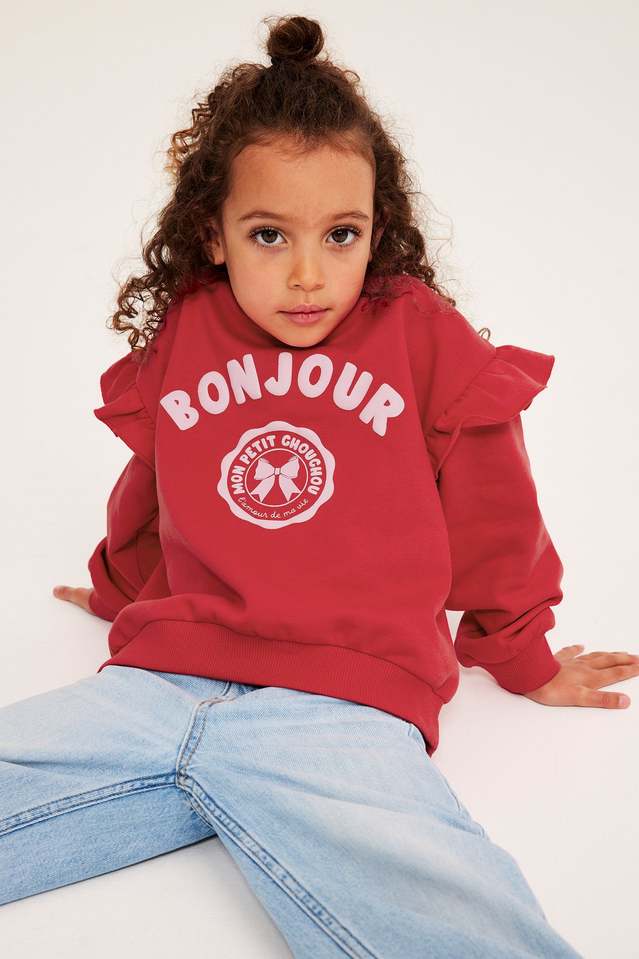 Framifrån: Ett barn i röd sweatshirt med volanger, "BONJOUR"-tryck och ljusblå löst sittande jeans.