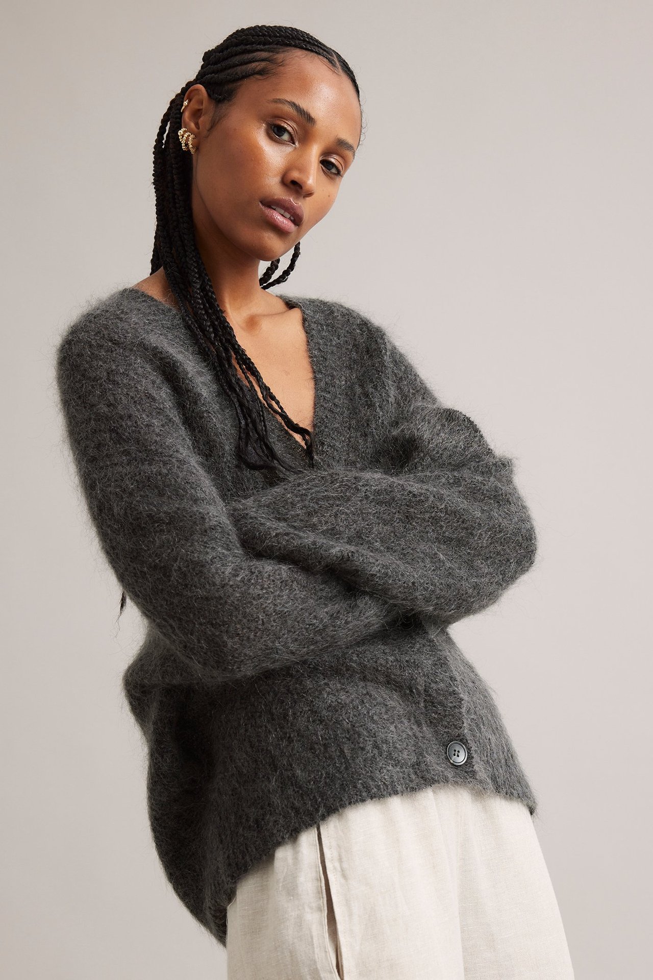 Frontvy av kvinna i grå mohairblandad kofta med V-ringning, ljusbeige byxor och guldfärgade örhängen.