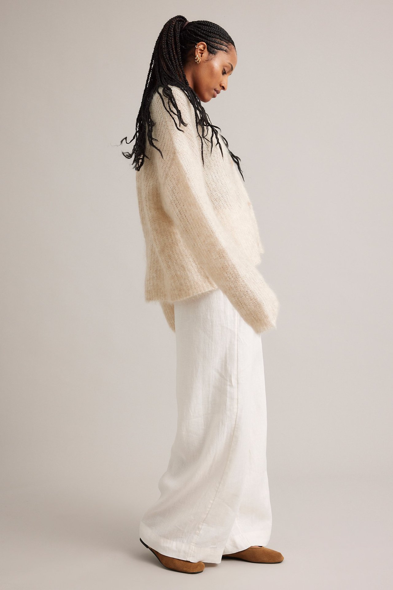 Sidovy av en kvinna i en ljusbeige, boxig kofta i mohairblandning, vita vida byxor och bruna skor.