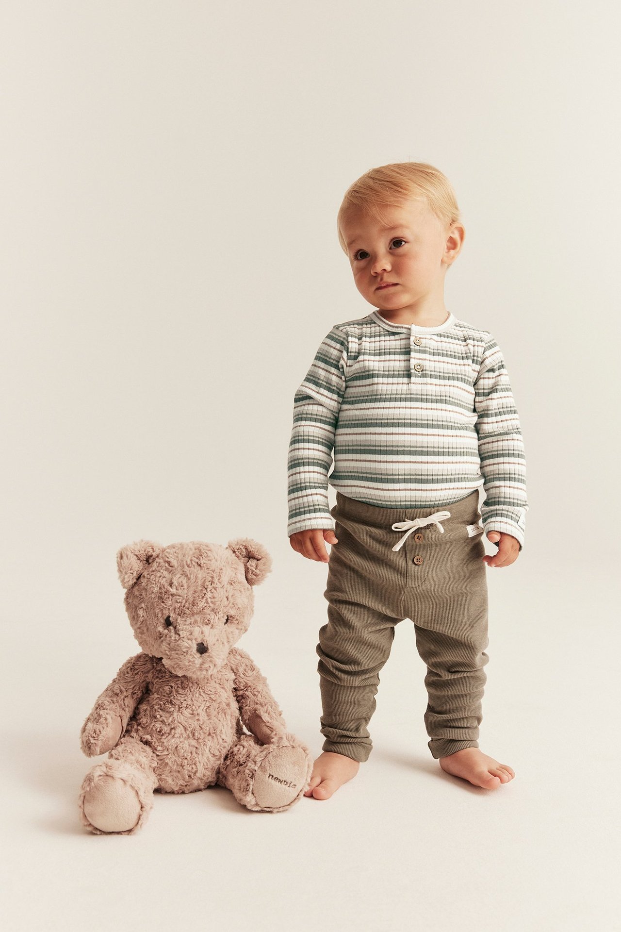 Vauva yllään luonnonvalkoinen ribattu raidallinen pitkähihainen body, oliivihousut ja ruskea nalle.
