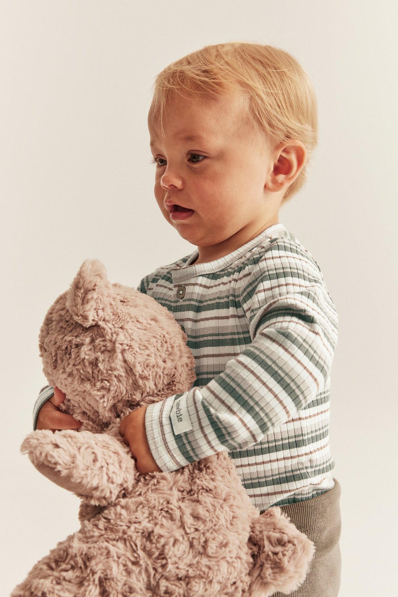 Lähikuva vauvasta yllään luonnonvalkoinen raidallinen pitkähihainen ribbipusero, pitäen ruskeaa nallea.