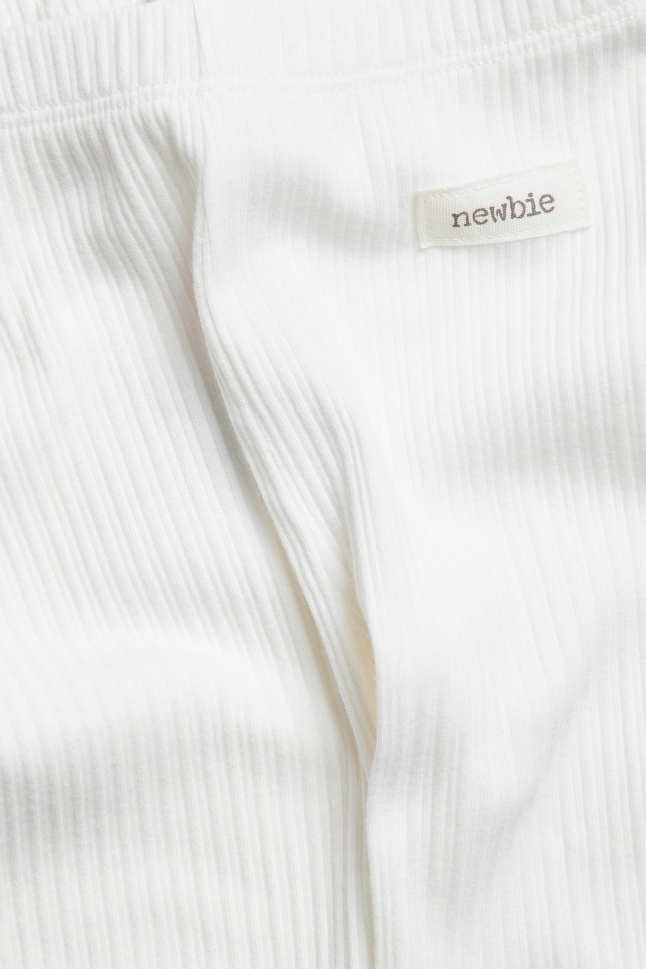 Zbliżenie na prążkowane legginsy niemowlęce w kolorze złamanej bieli z gumką w pasie i metką Newbie.