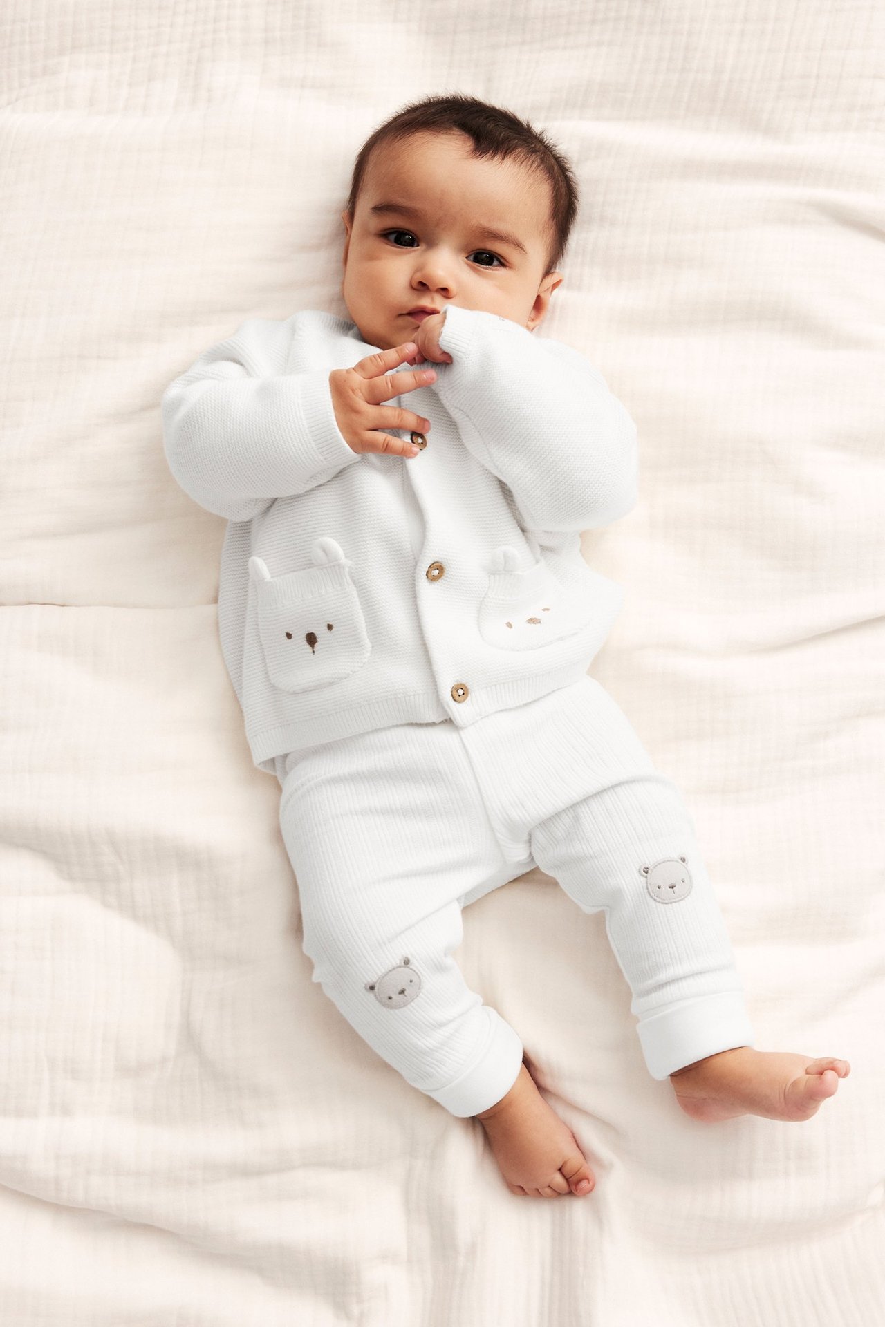 Ovenfra: Baby i offwhite strikket bomullskofta med broderte bjørnelommer og matchende ribbebukser.